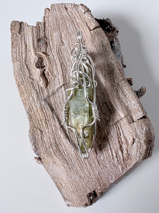 silver wire wrapped green kyanite pendant on a piece of driftwood against a white background