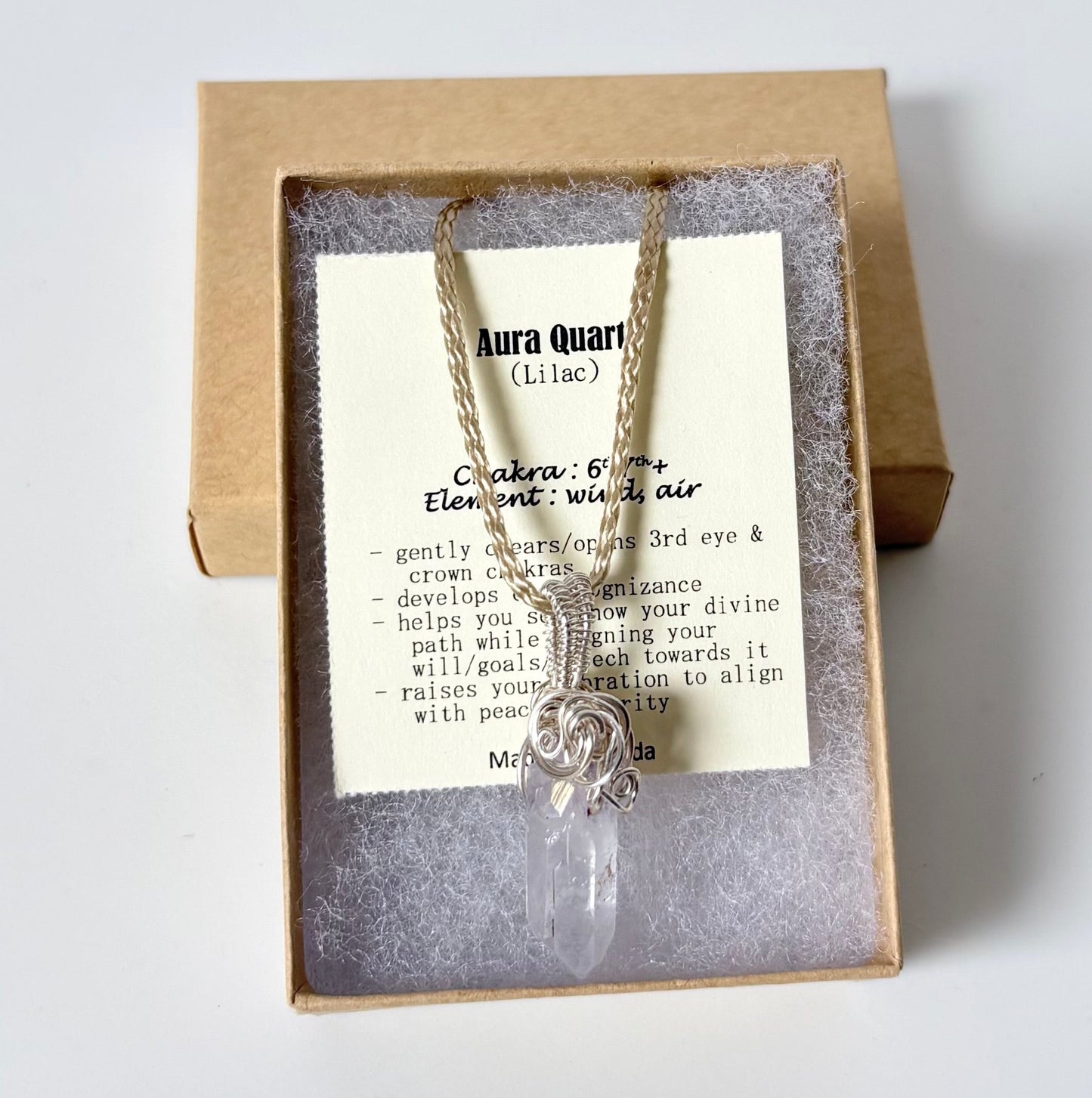 Necklace with a silver wire wrapped Aura Quartz (lilac) pendant on an information card with text explaining the folklore and metaphysical properties placed in a brown paper kraft jewelry box for gift giving all on a white surface.