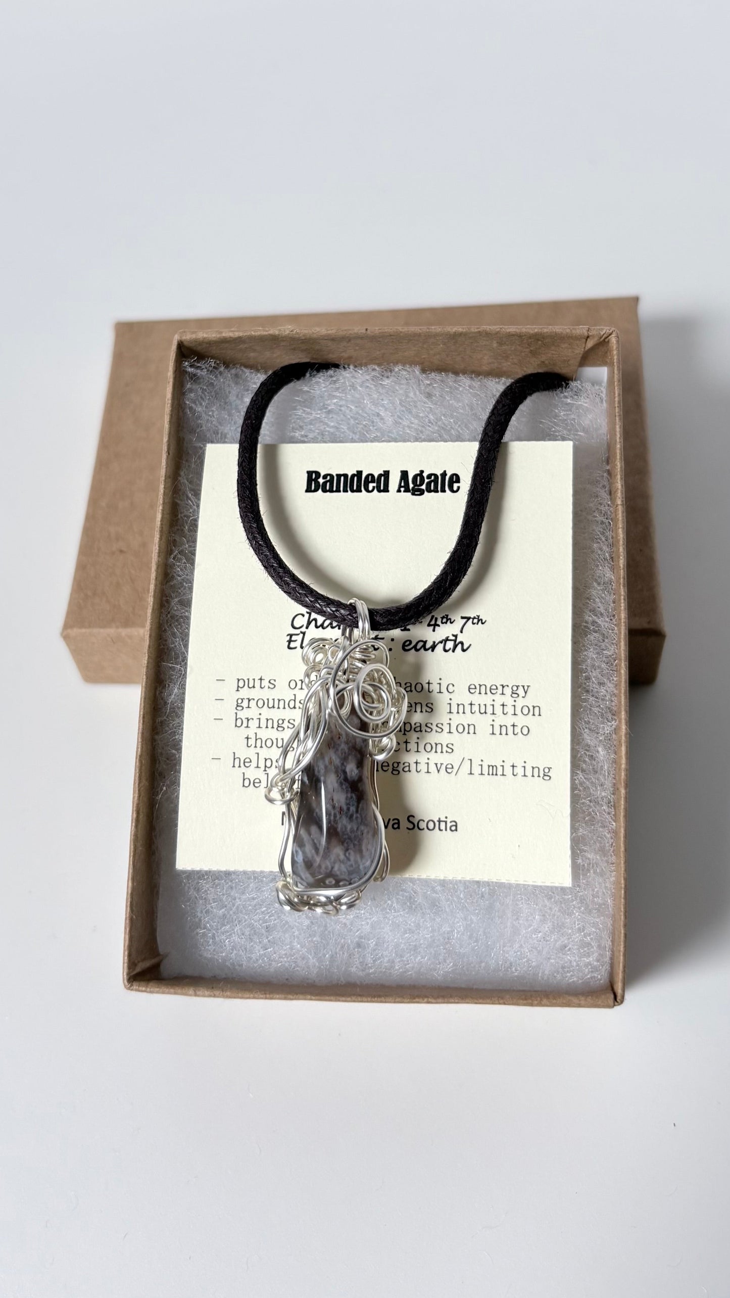 Necklace with a silver wire wrapped banded agate pendant on an information card with text explaining the folklore and metaphysical properties placed in a brown paper kraft jewelry box for gift giving all on a white surface.