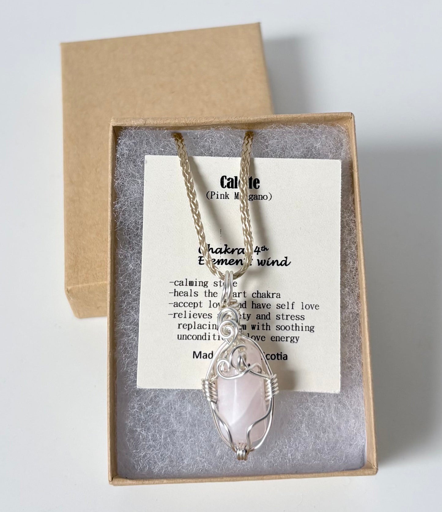 Necklace with a silver wire wrapped calcite (pink mangano) pendant on an information card with text explaining the folklore and metaphysical properties placed in a brown paper kraft jewelry box for gift giving all on a white surface.