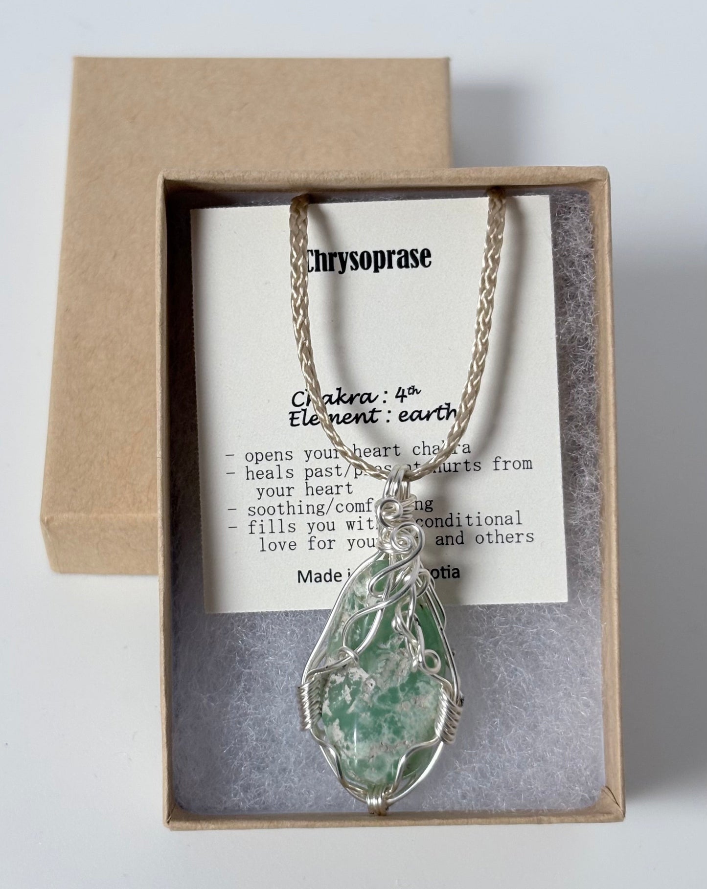 Necklace with a silver wire wrapped chrysoprase pendant on an information card with text explaining the folklore and metaphysical properties placed in a brown paper kraft jewelry box for gift giving all on a white surface.