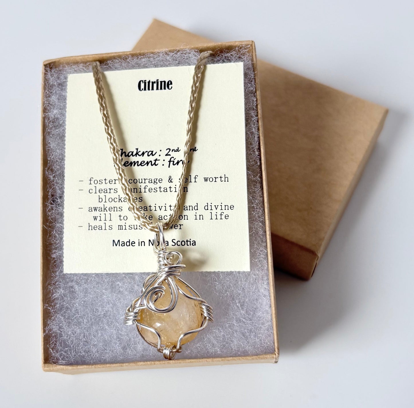 Necklace with a silver wire wrapped citrine pendant on an information card with text explaining the folklore and metaphysical properties placed in a brown paper kraft jewelry box for gift giving all on a white surface.