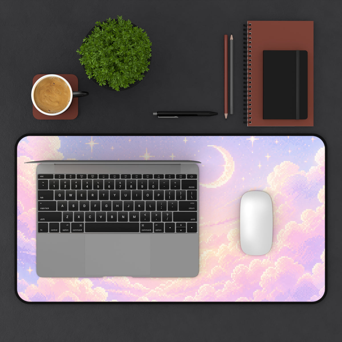 12 by 22 inch pastel celestial desk mat shown on a dark desktop with a laptop and white mouse for size reference. The large keyboard mat features a soft pixel-art sky design with pink and lavender clouds, glowing stars and a crescent moon, finished with rounded corners and a black border. Coffee, a green plant, pens, pencils, a spiral notebook and a small black journal are arranged around the mat to show how it fits in a workspace.