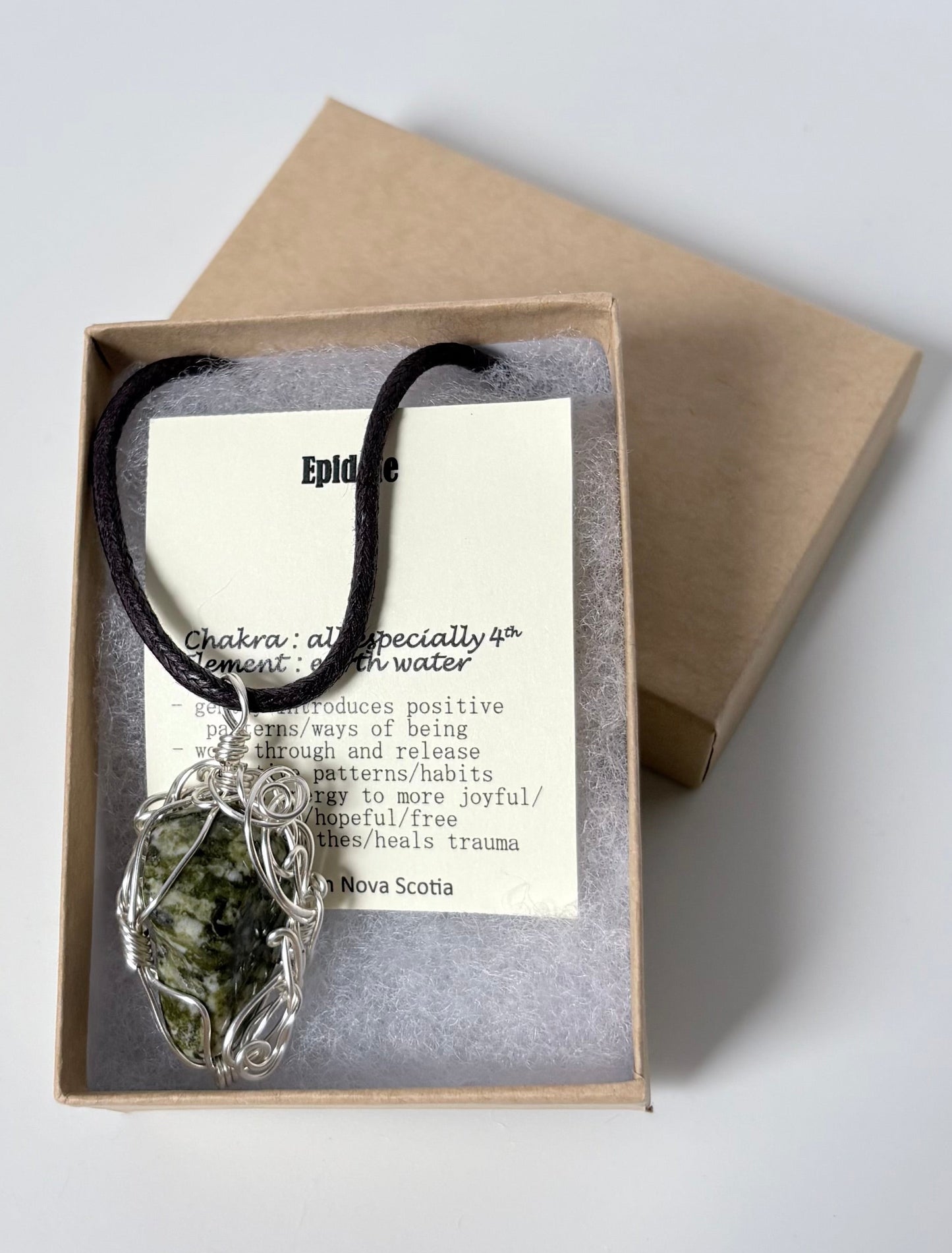 Necklace with a silver wire wrapped epidot pendant on an information card with text explaining the folklore and metaphysical properties placed in a brown paper kraft jewelry box for gift giving all on a white surface.