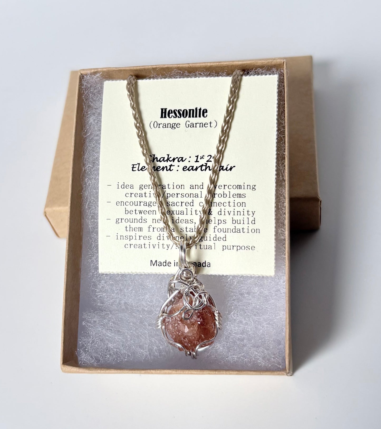 Necklace with a silver wire wrapped hessonite pendant on an information card with text explaining the folklore and metaphysical properties placed in a brown paper kraft jewelry box for gift giving all on a white surface.