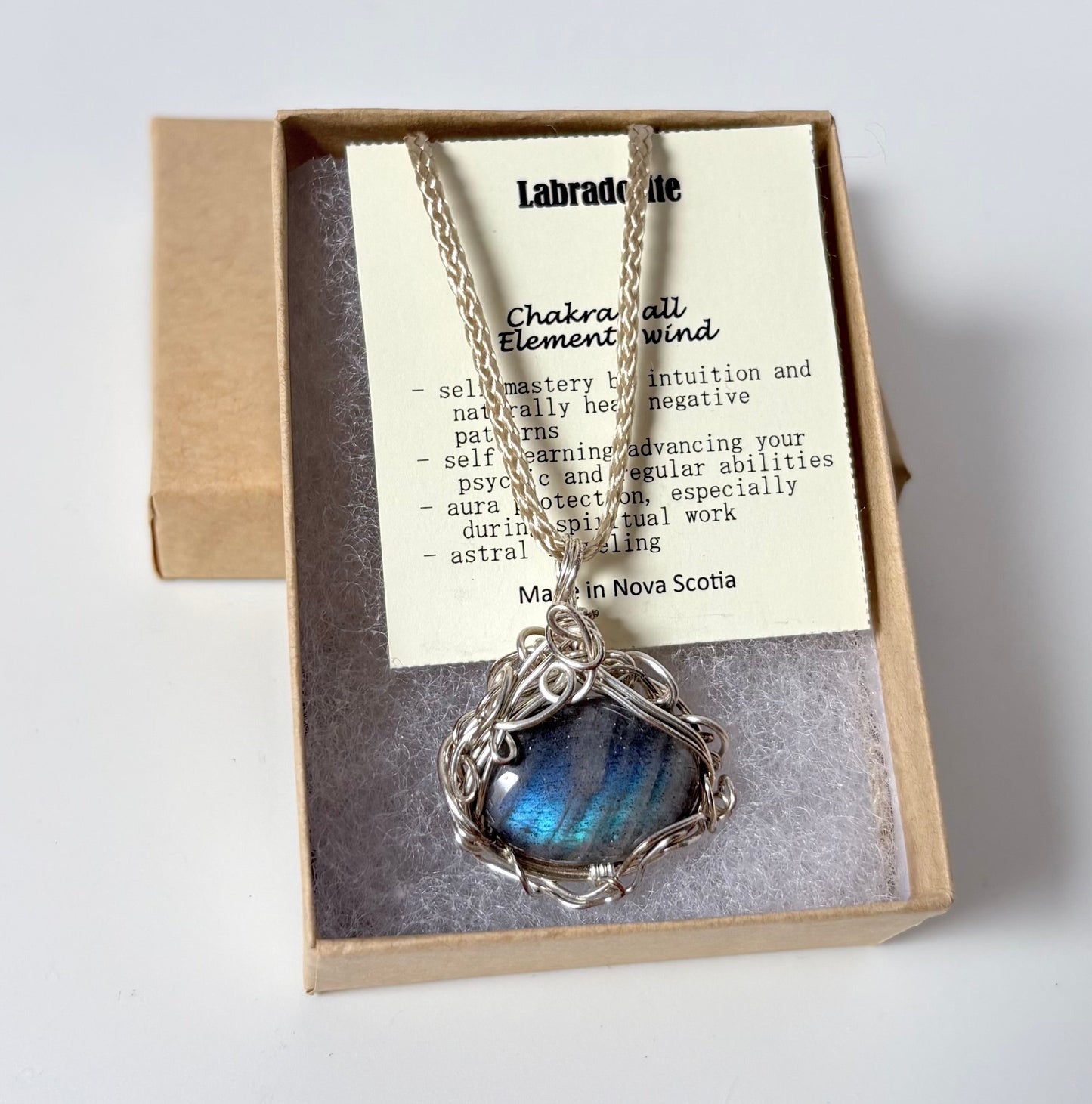 Necklace with a silver wire wrapped labradorite pendant on an information card with text explaining the folklore and metaphysical properties placed in a brown paper kraft jewelry box for gift giving all on a white surface.