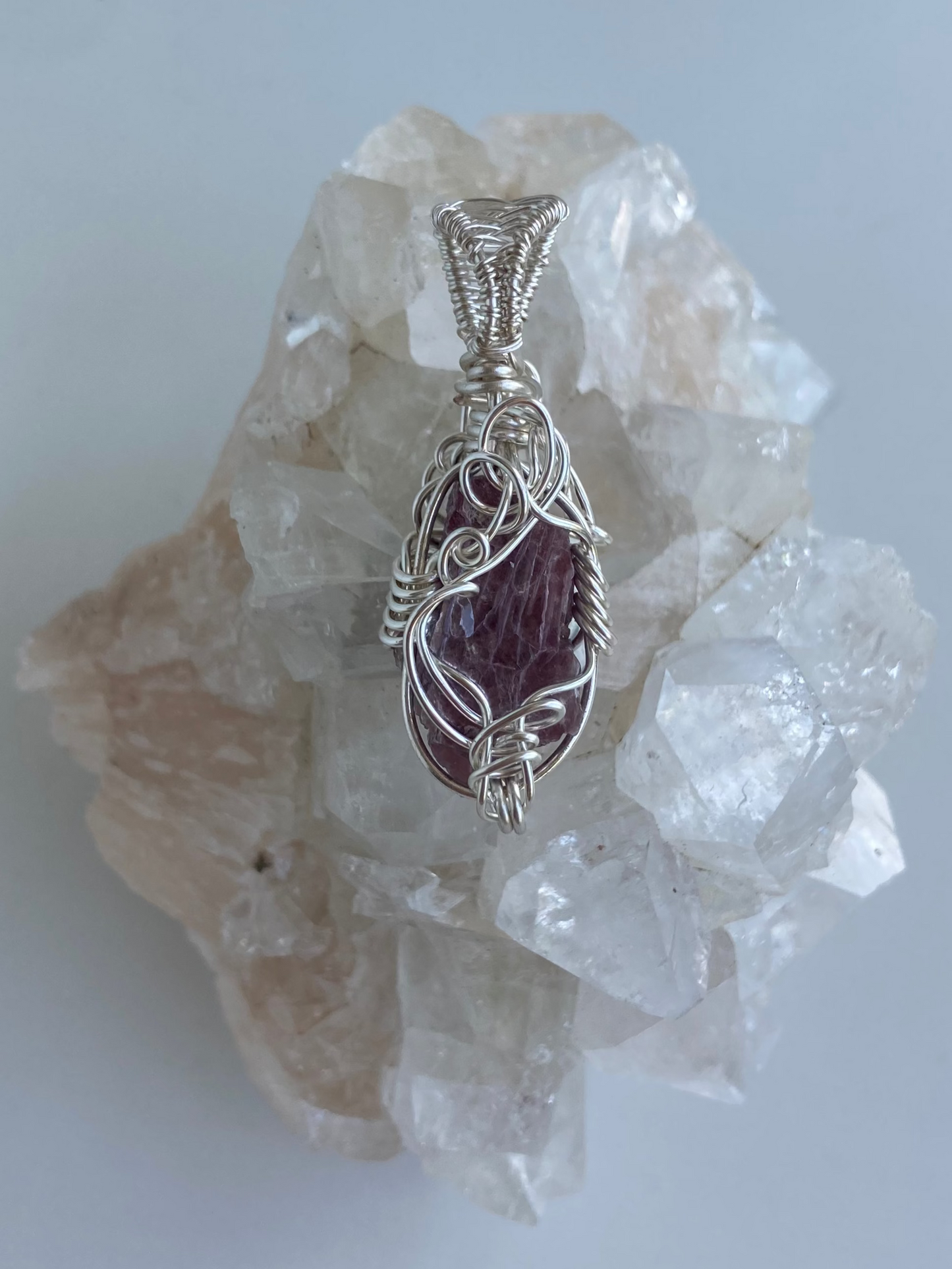 Lepidolite Pendant – Front view of a wire-wrapped purple lepidolite stone pendant, handcrafted in silver-plated copper wire with a unique spiral design — photographed on a clear apophyllite crystal cluster with a white background.
