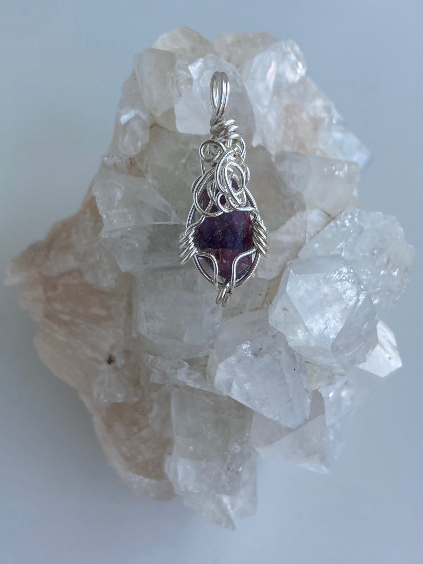 Lepidolite Pendant – Front view of a wire-wrapped purple lepidolite stone pendant, handcrafted in silver-plated copper wire with a unique spiral design — photographed on a clear apophyllite crystal cluster surface.