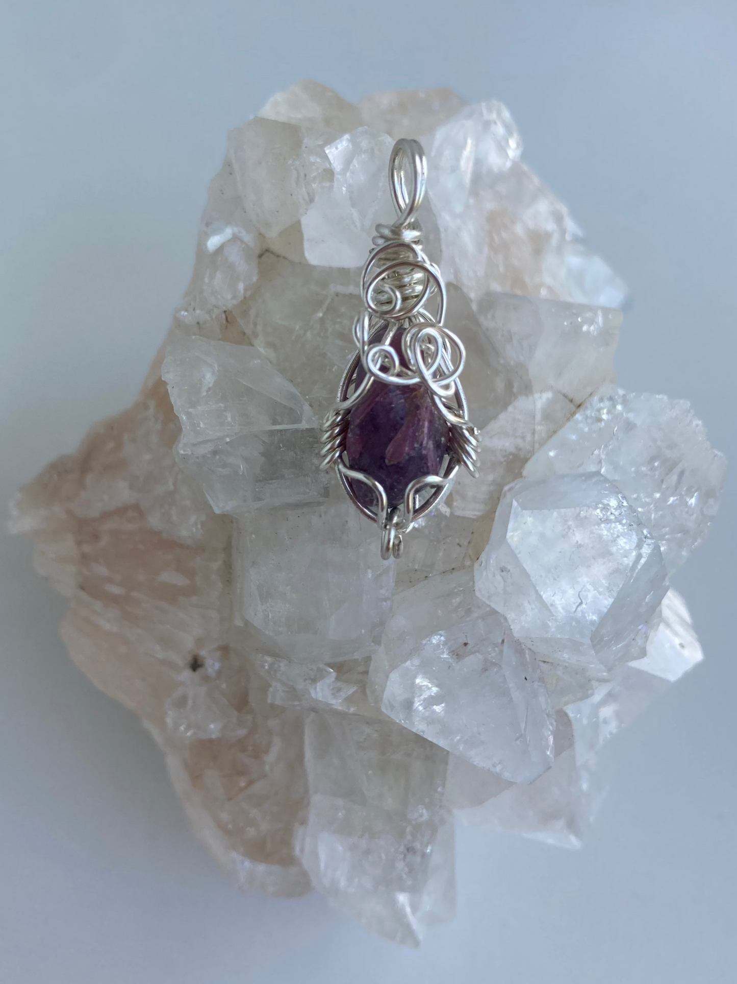 Lepidolite Pendant – Back view of a wire-wrapped purple lepidolite stone pendant, handcrafted in silver-plated copper wire with a unique spiral design — photographed on a clear apophyllite crystal cluster surface.