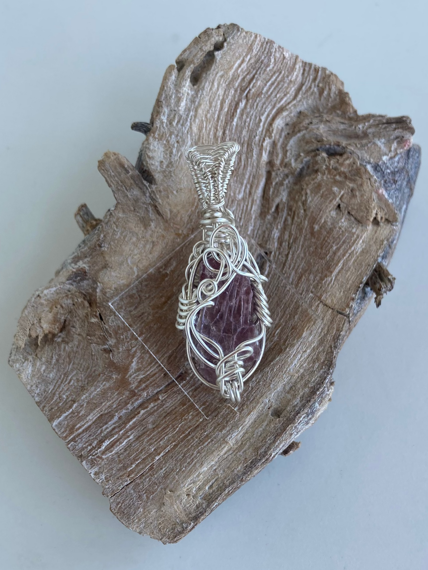 Lepidolite Pendant – Front view of a wire-wrapped purple lepidolite stone pendant, handcrafted in silver-plated copper wire with a unique spiral design — photographed on a wood surface with a white background.