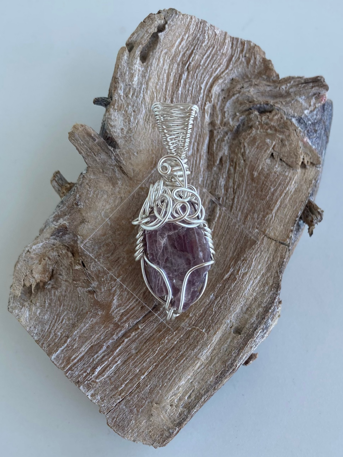 Lepidolite Pendant – Back view of a wire-wrapped purple lepidolite stone pendant, handcrafted in silver-plated copper wire with a unique spiral design — photographed on a wood surface with a white background.