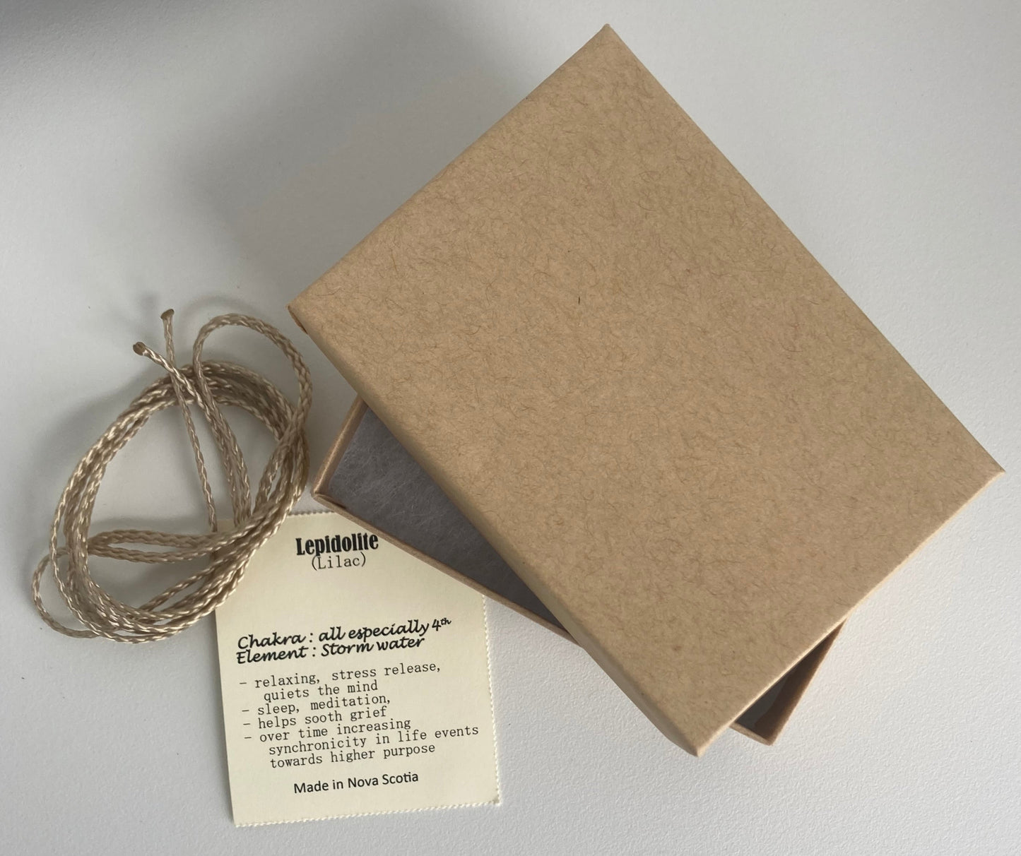 Lepidolite pendant packaging set — includes a metallic champagne-colored nylon cord to wear your pendant, a cream-colored information card listing the stone type along with its associated chakra(s), element(s), and metaphysical properties, and a light brown kraft paper jewelry box with white padding inside to hold the pendant, cord, and card — great for gift giving. Photographed on a white background.