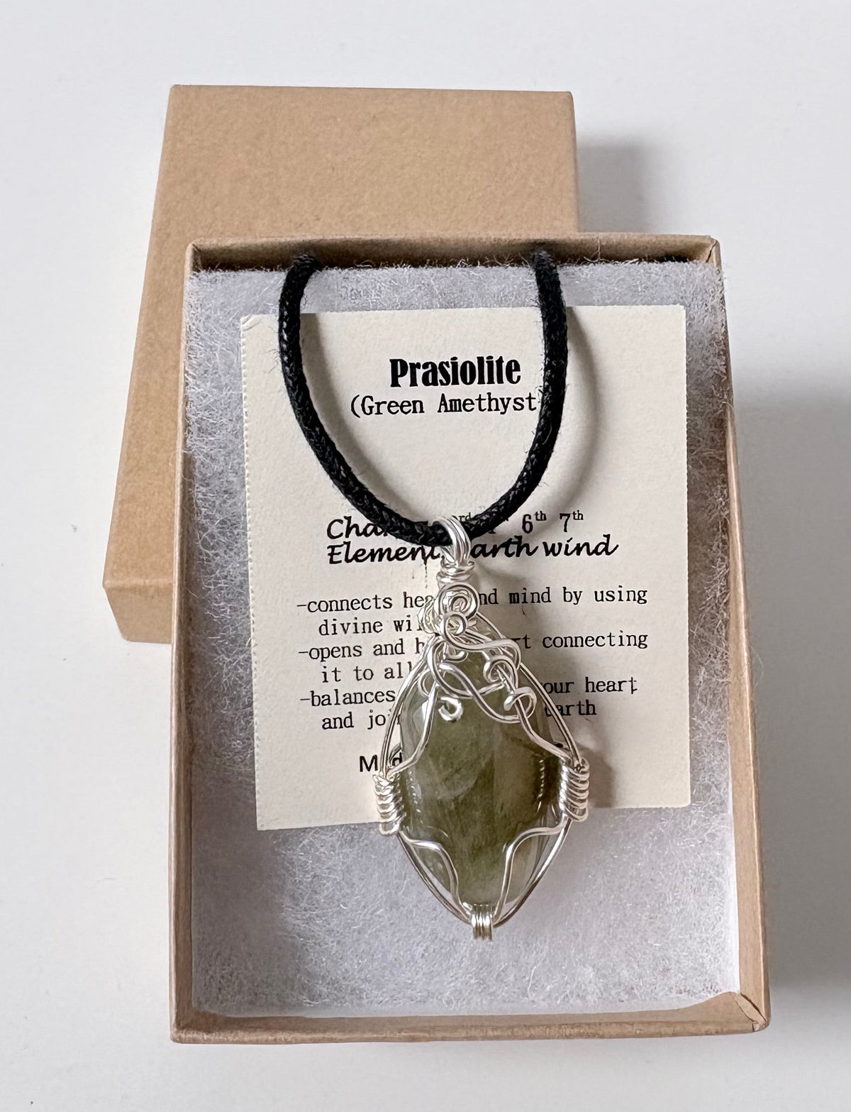 Necklace with a silver wire wrapped prasiolite (green amethyst) pendant on an information card with text explaining the folklore and metaphysical properties placed in a brown paper kraft jewelry box for gift giving all on a white surface.