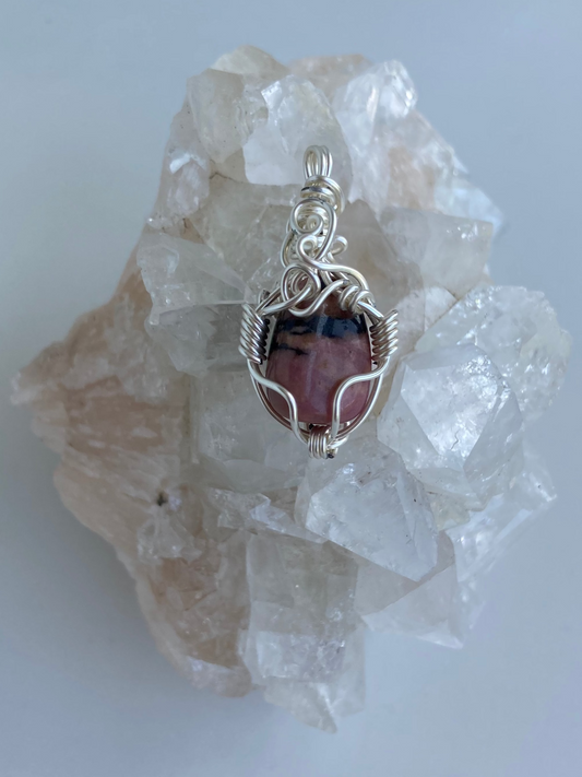 Rhodonite Pendant – Back view of a wire-wrapped pink and black rhodonite stone pendant, handcrafted in silver-plated copper wire with a unique spiral design — photographed on a clear apophyllite crystal cluster surface with a white background.