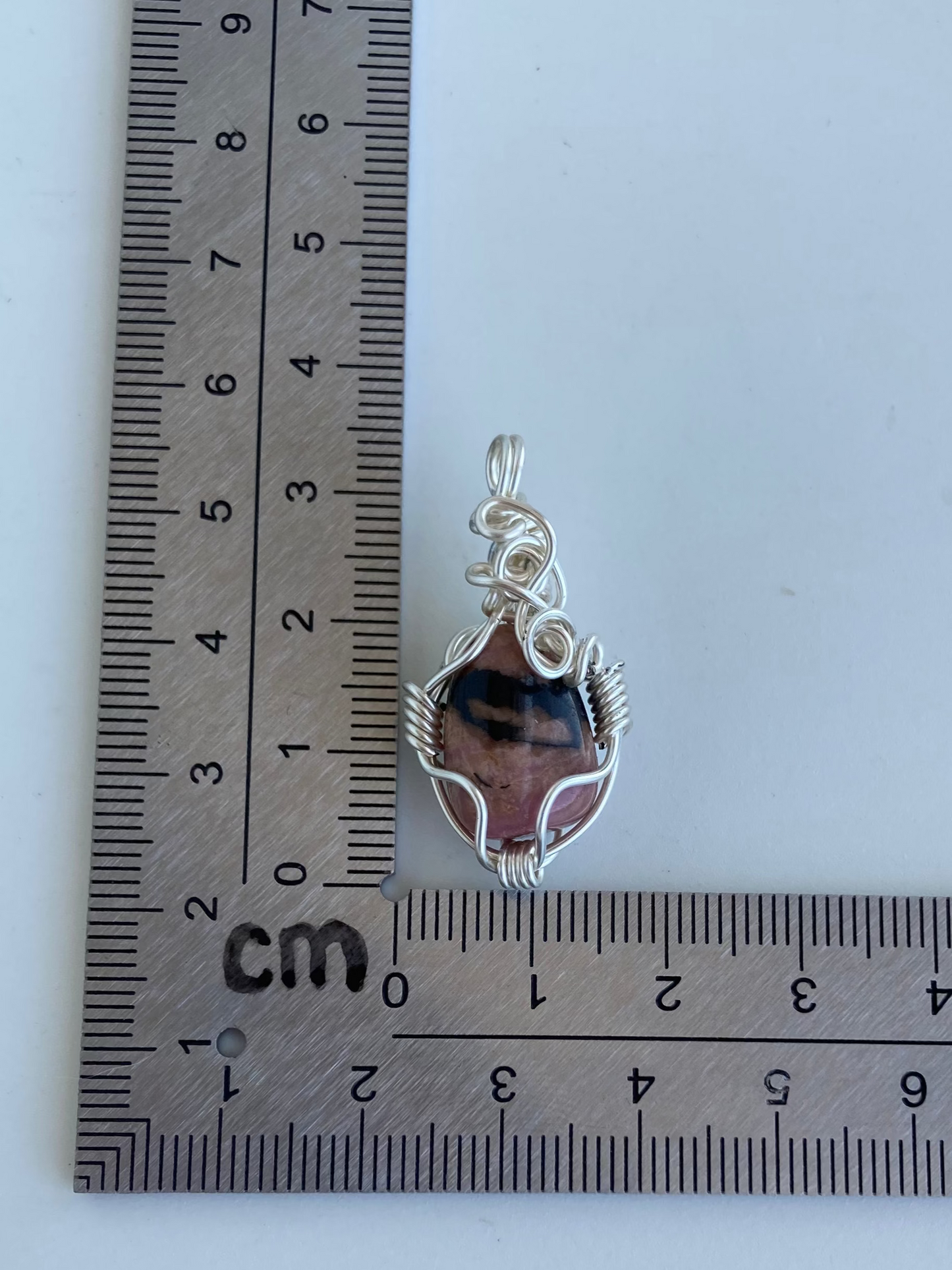 Rhodonite Pendant – Front view of a wire-wrapped pink and black rhodonite stone pendant shown beside an L-shaped centimeter ruler for size reference. Handcrafted in silver-plated copper wire with a unique spiral design — photographed on a white surface.
