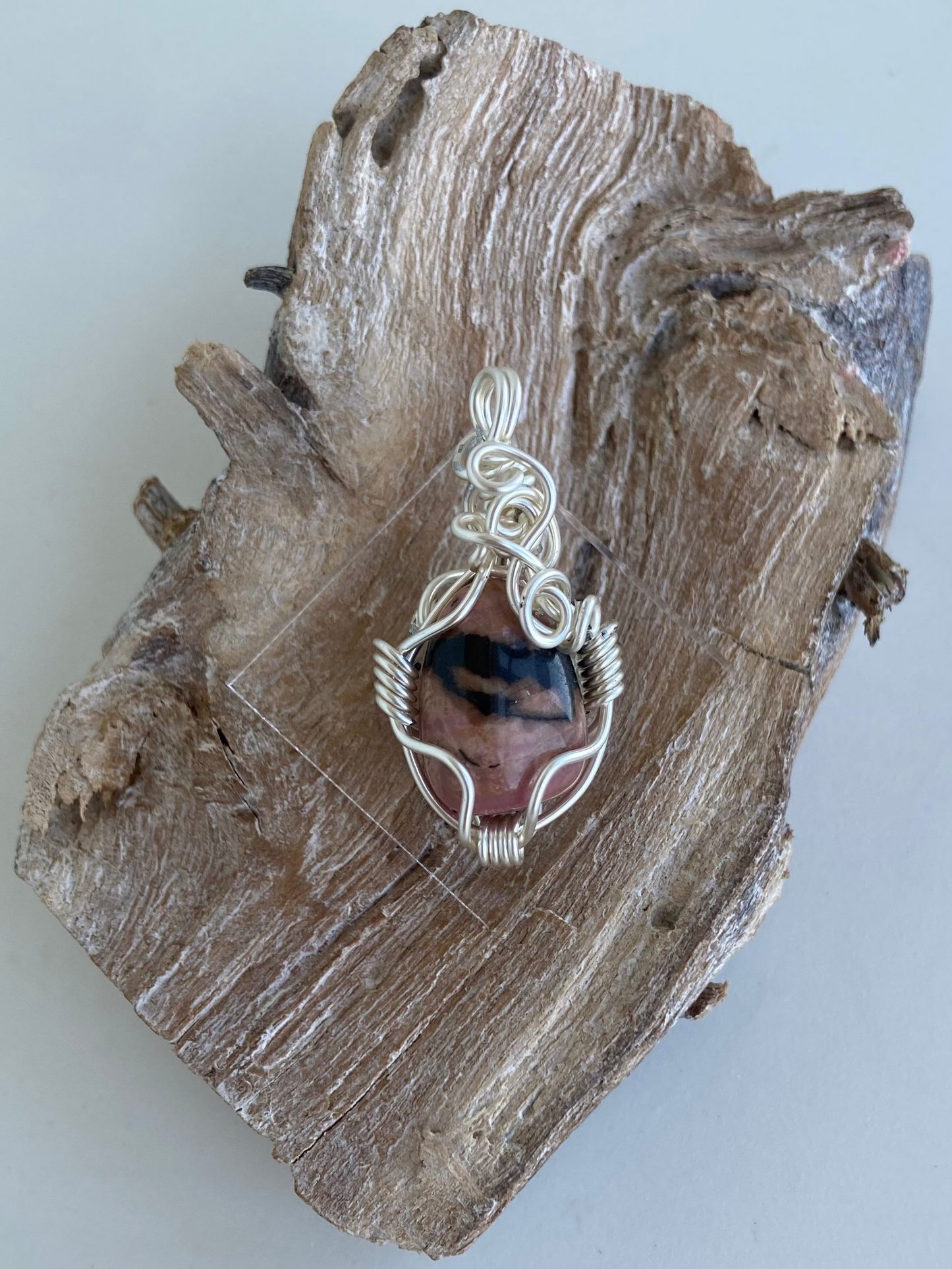 Rhodonite Pendant – Front view of a wire-wrapped pink and black rhodonite stone pendant, handcrafted in silver-plated copper wire with a unique spiral design — photographed on a wood surface with a white background.