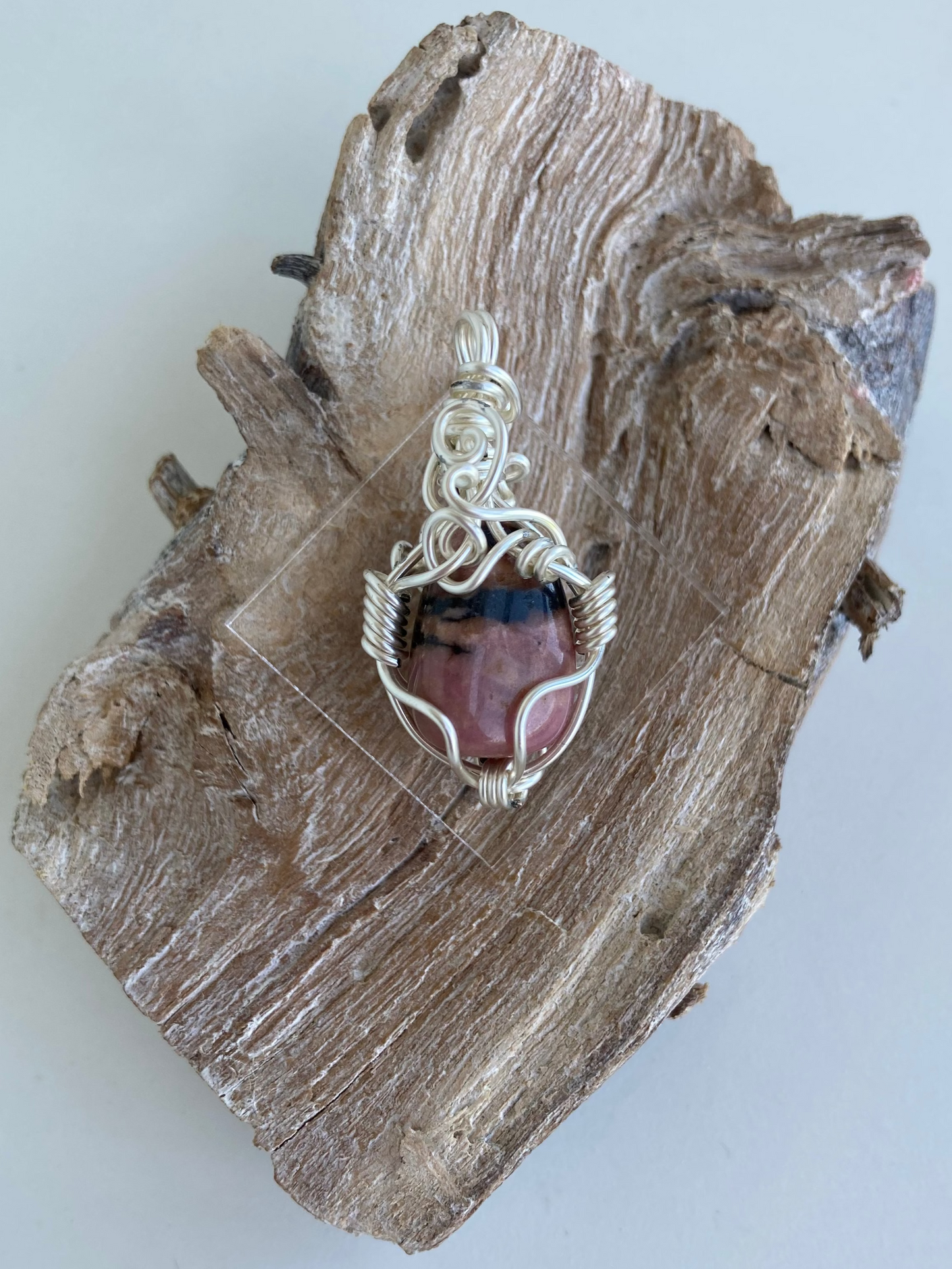 Rhodonite Pendant – Back view of a wire-wrapped pink and black rhodonite stone pendant, handcrafted in silver-plated copper wire with a unique spiral design — photographed on a wood surface with a white background.