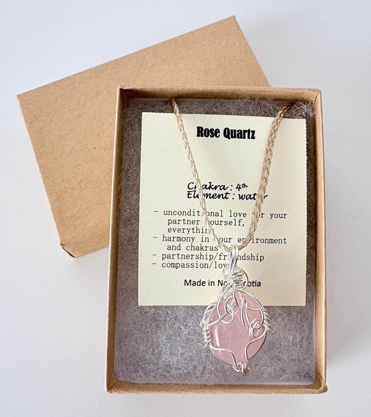 Necklace with a silver wire wrapped rose quartz pendant on an information card with text explaining the folklore and metaphysical properties placed in a brown paper kraft jewelry box for gift giving all on a white surface.
