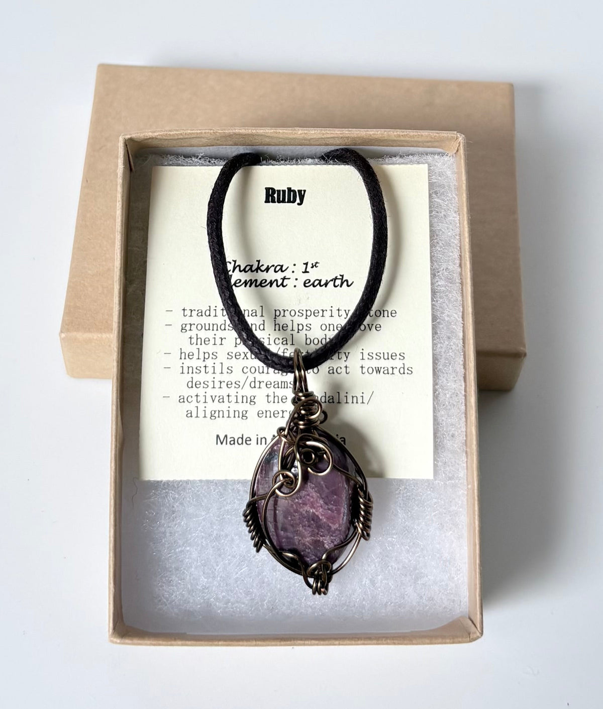 Necklace with an antique brass wire wrapped ruby pendant on an information card with text explaining the folklore and metaphysical properties placed in a brown paper kraft jewelry box for gift giving all on a white surface.