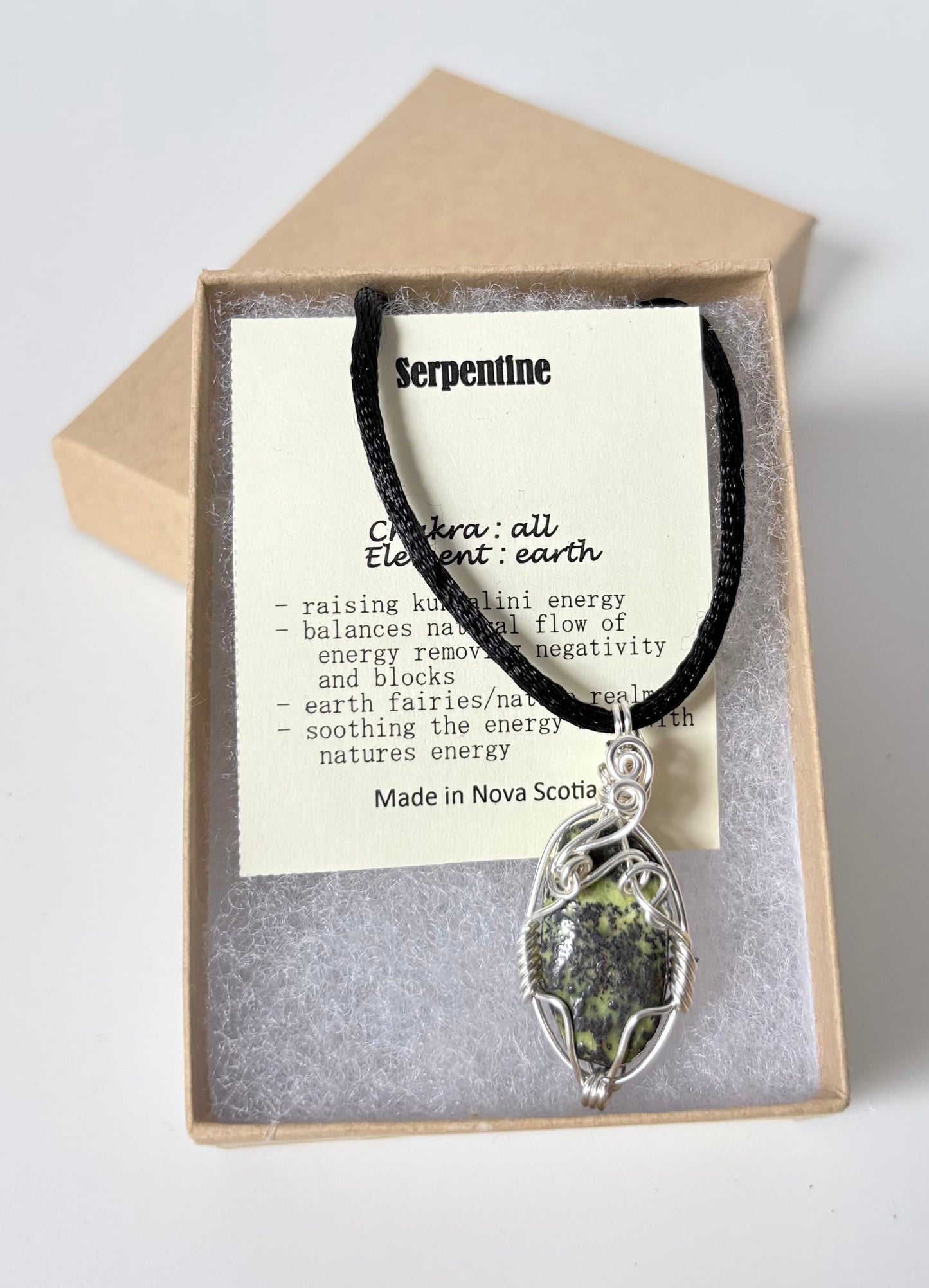 Necklace with a silver wire wrapped serpentine pendant on an information card with text explaining the folklore and metaphysical properties placed in a brown paper kraft jewelry box for gift giving all on a white surface.