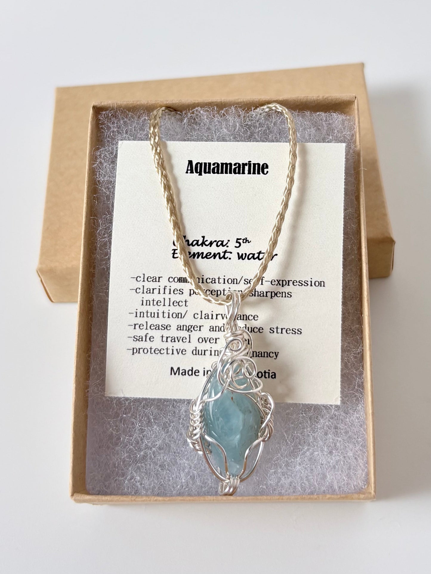 Necklace with a silver wire wrapped aquamarine pendant on an information card with text explaining the folklore and metaphysical properties placed in a brown paper kraft jewelry box for gift giving all on a white surface.