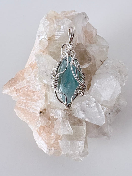 silver wire wrapped aquamarine pendant displayed on a clear apophyllite cluster all on a white surface.