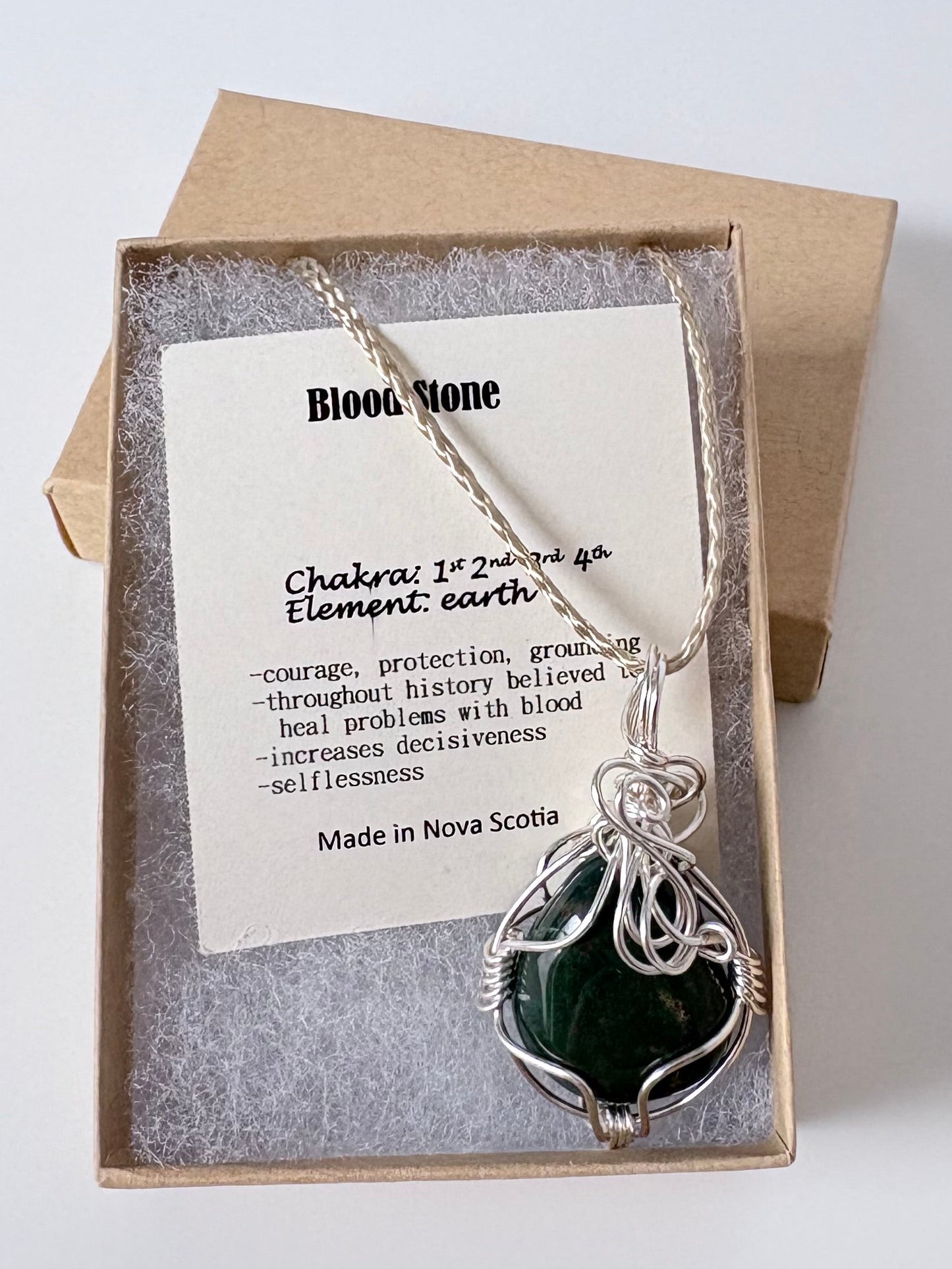 Necklace with a silver wire wrapped bloodstone pendant on an information card with text explaining the folklore and metaphysical properties placed in a brown paper kraft jewelry box for gift giving all on a white surface.
