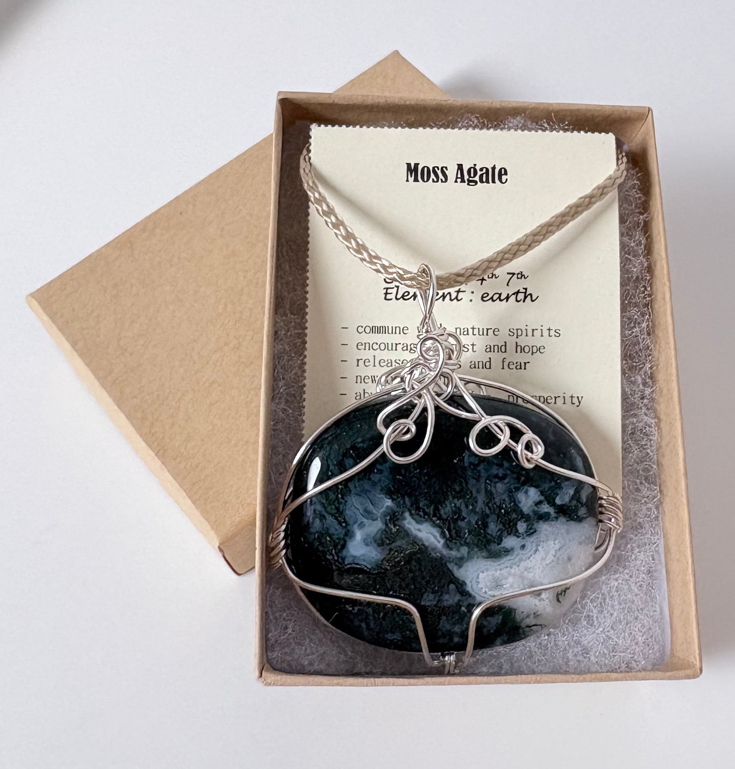 Necklace with a silver wire wrapped moss agate pendant on an information card with text explaining the folklore and metaphysical properties placed in a brown paper kraft jewelry box for gift giving all on a white surface.