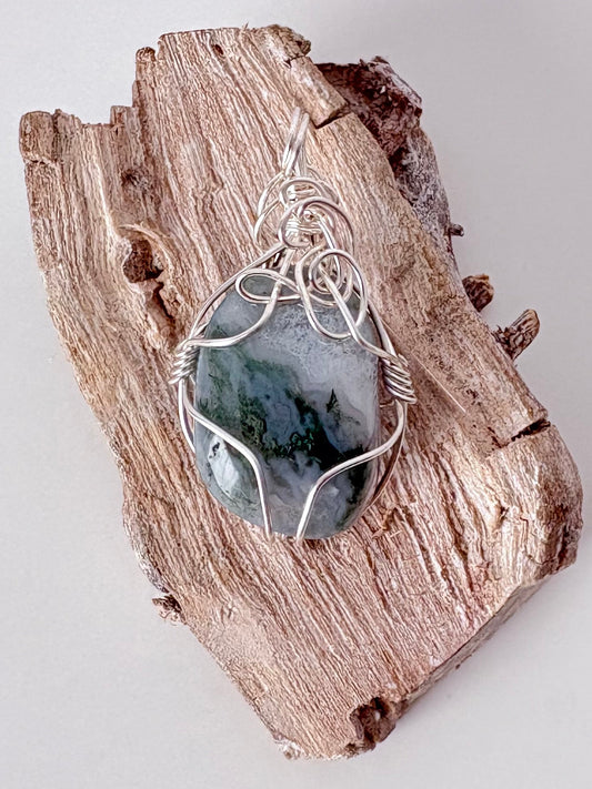 Silver wire-wrapped moss agate pendant on a piece of driftwood against a light background