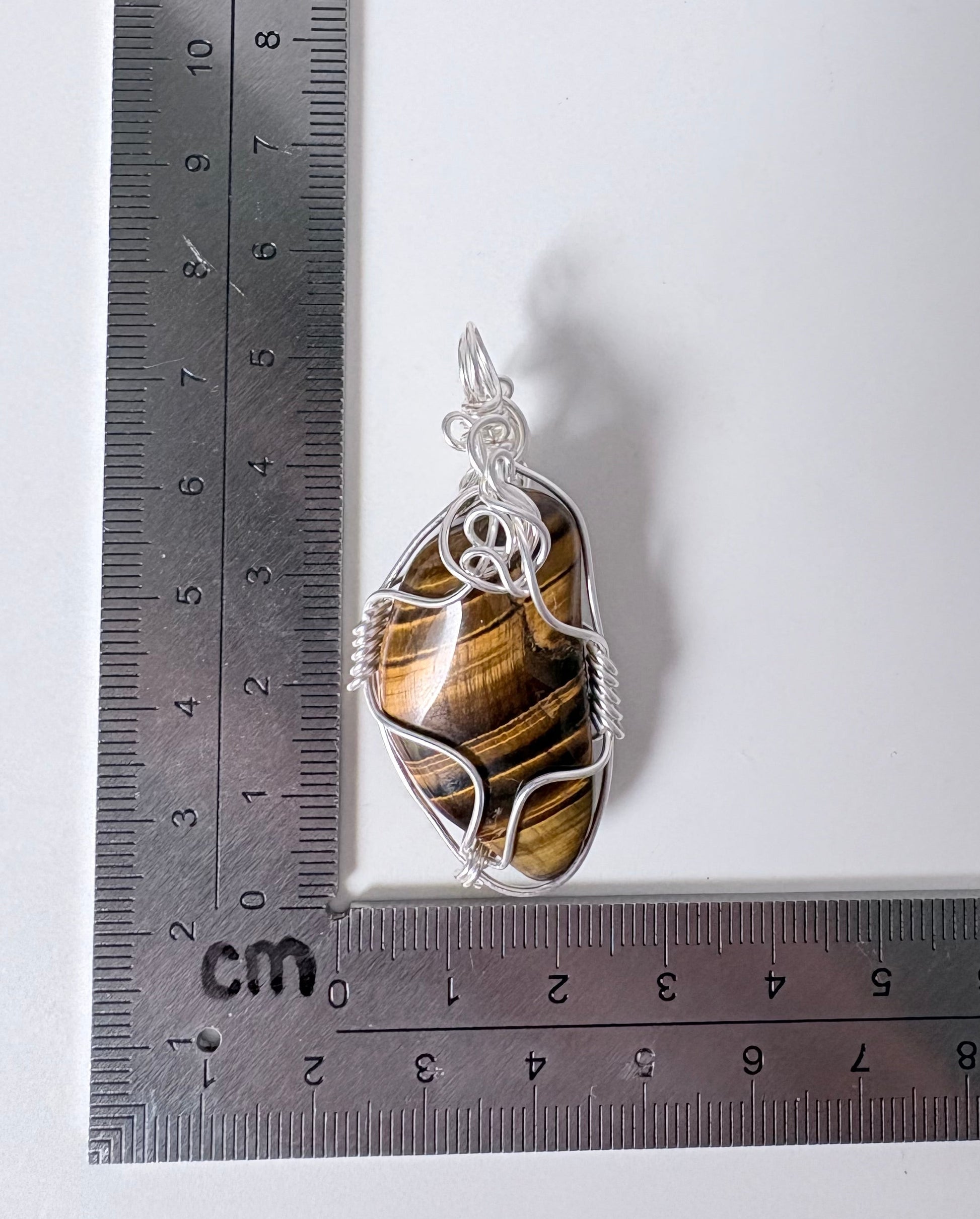Pendant with tiger eye stone and silver wire wrapping on a white background with a ruler for scale.