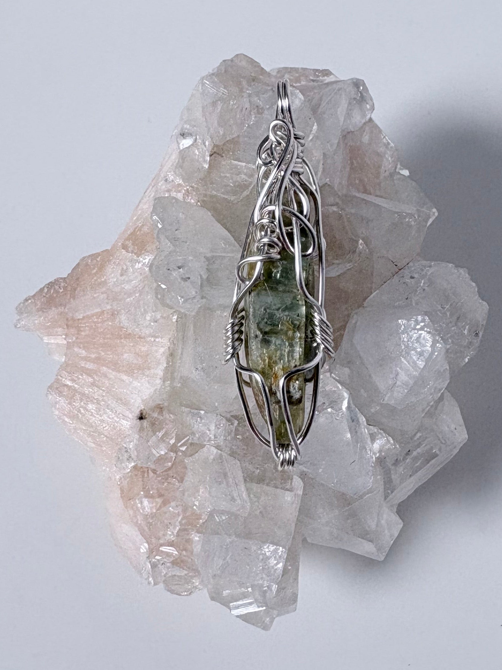 silver wire wrapped green kyanite pendant on clear crystal cluster with a white background