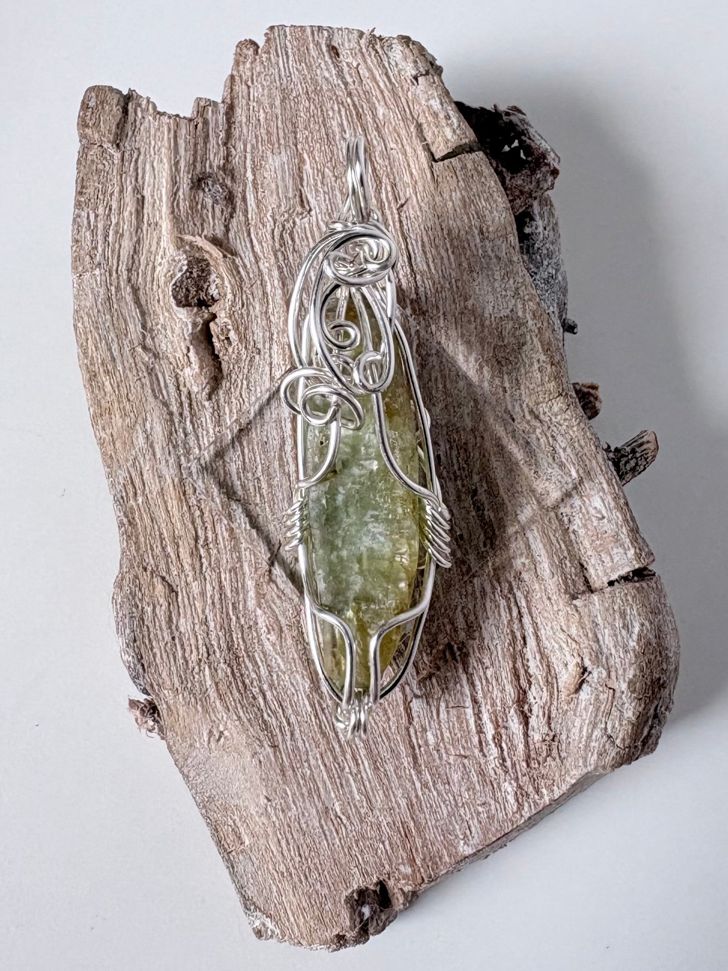 silver wire wrapped green kyanite pendant on a piece of driftwood against a white background