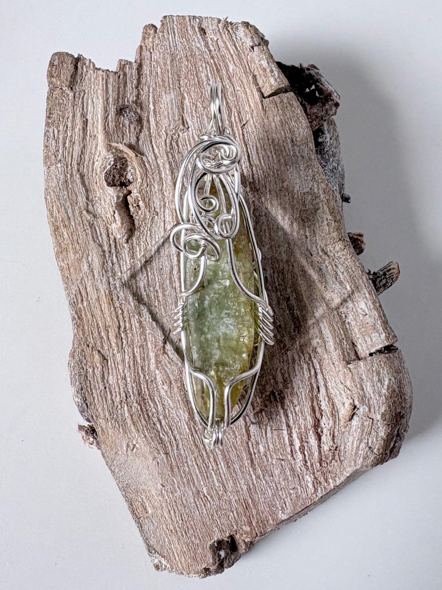 silver wire wrapped green kyanite pendant on a piece of driftwood against a white background