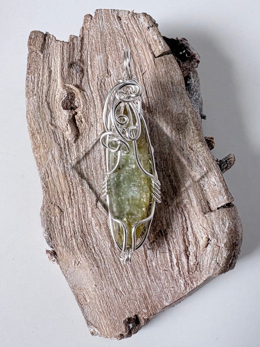silver wire wrapped green kyanite pendant on a piece of driftwood against a white background