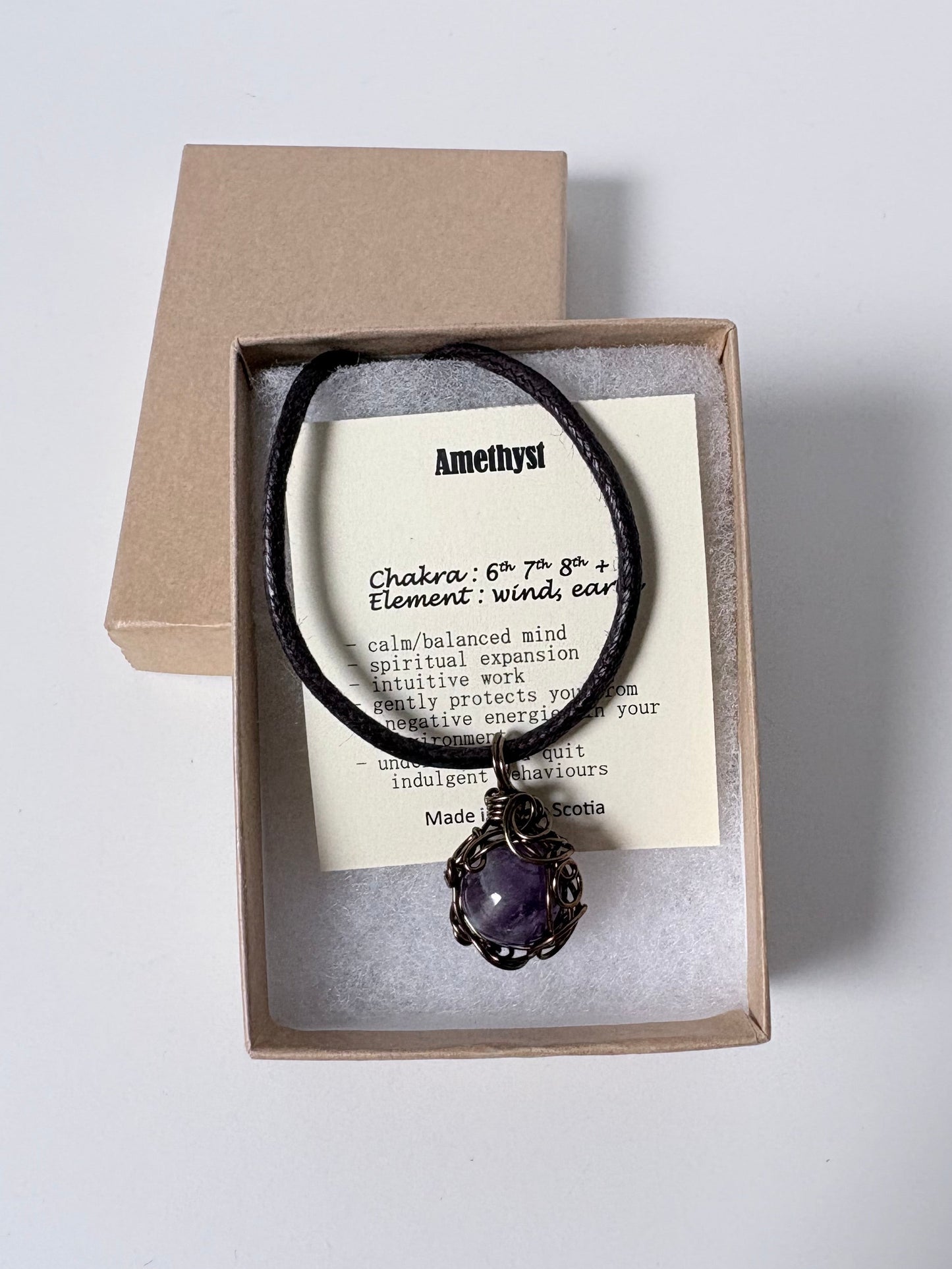 Necklace with an antique brass wire wrapped amethyst pendant on an information card with text explaining the folklore and metaphysical properties placed a brown paper kraft jewelry box for gift giving on a white surface.