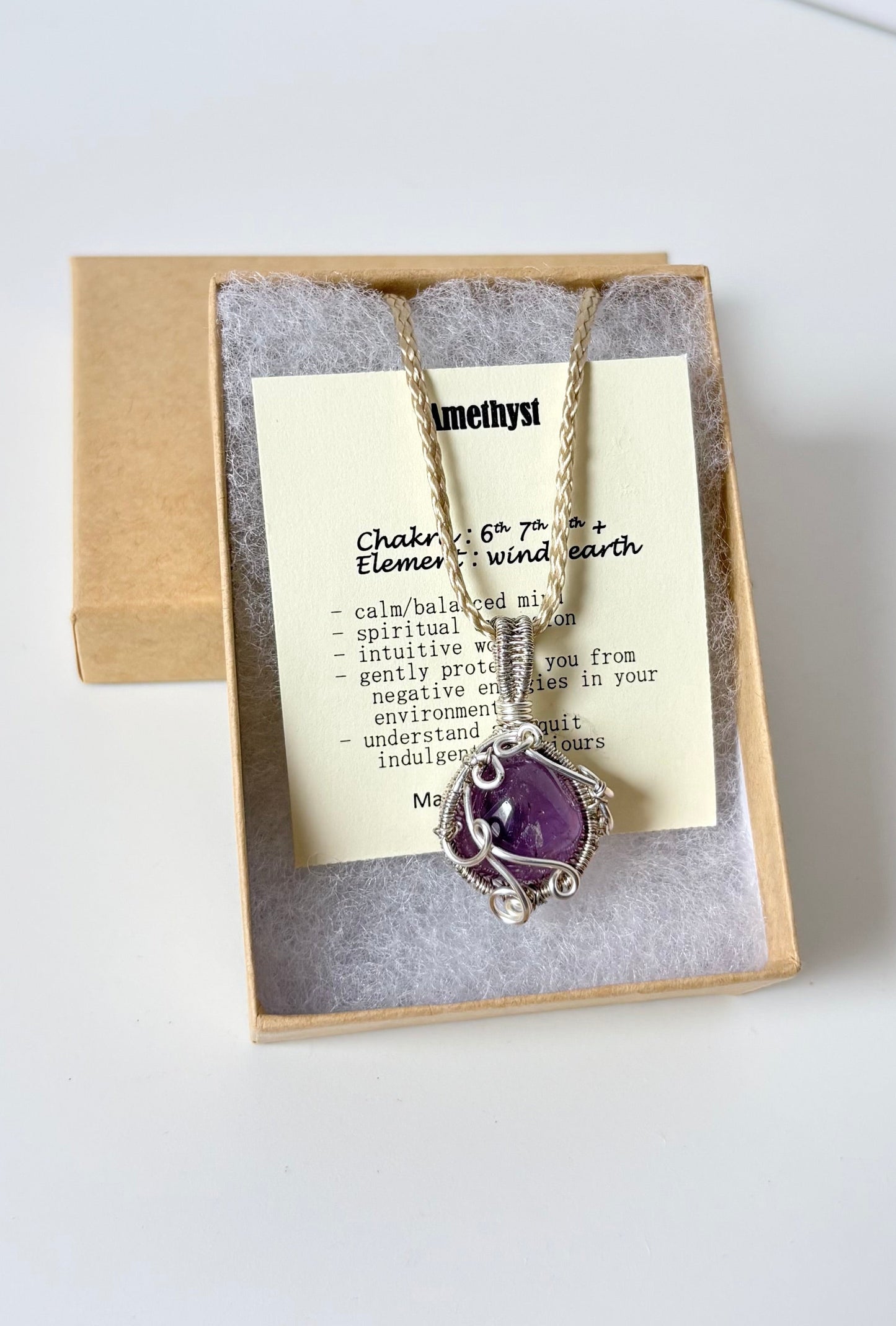 Necklace with an amethyst pendant on an information card with text explaining the folklore and metaphysical properties placed a brown paper kraft jewelry box for gift giving on a white surface.