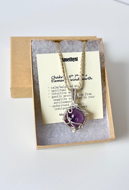 Necklace with an amethyst pendant on an information card with text explaining the folklore and metaphysical properties placed a brown paper kraft jewelry box for gift giving on a white surface.