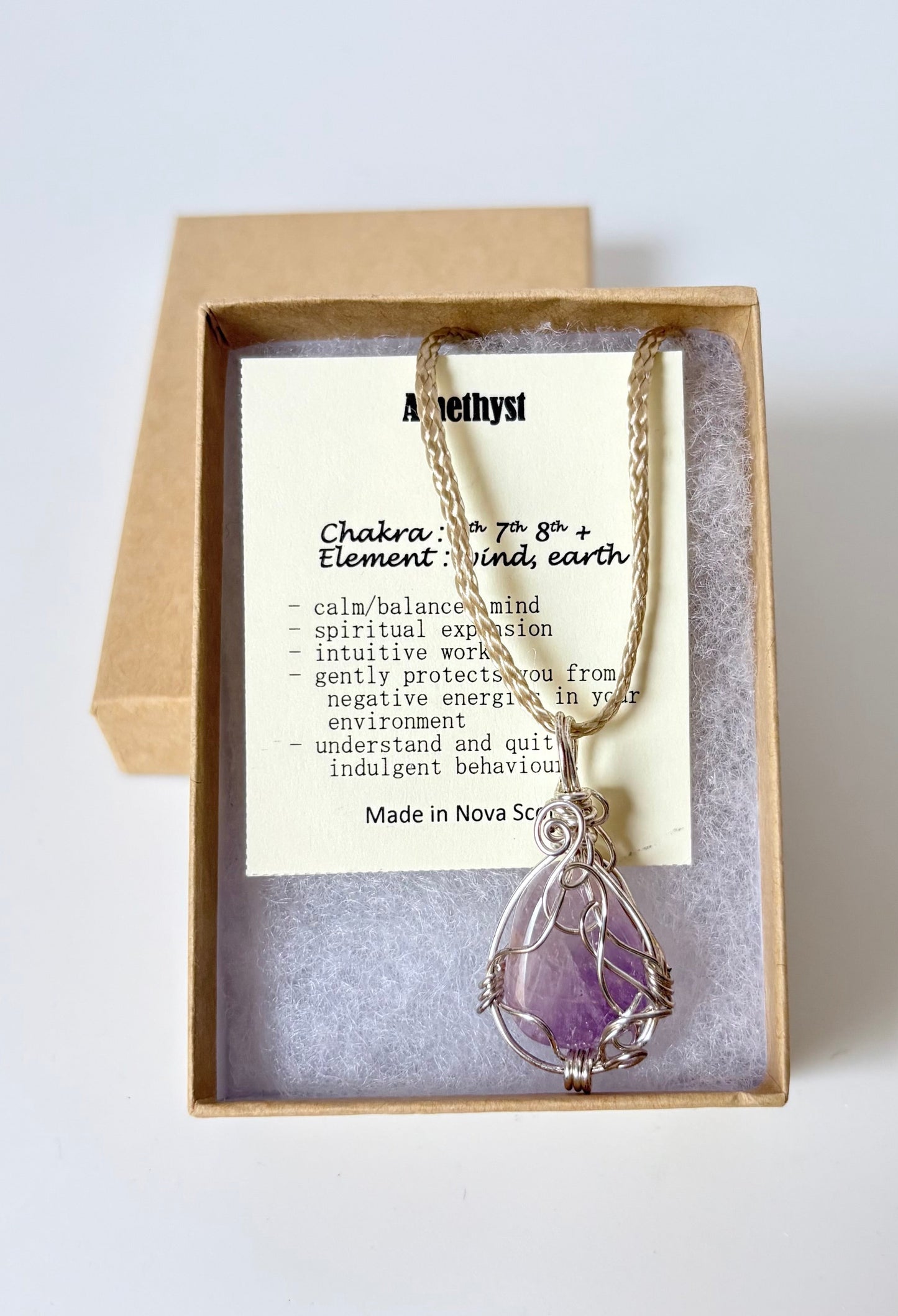 Necklace with a silver wire wrapped amethyst pendant on an information card with text explaining the folklore and metaphysical properties placed on a brown paper kraft jewelry box for gift giving on a white surface.