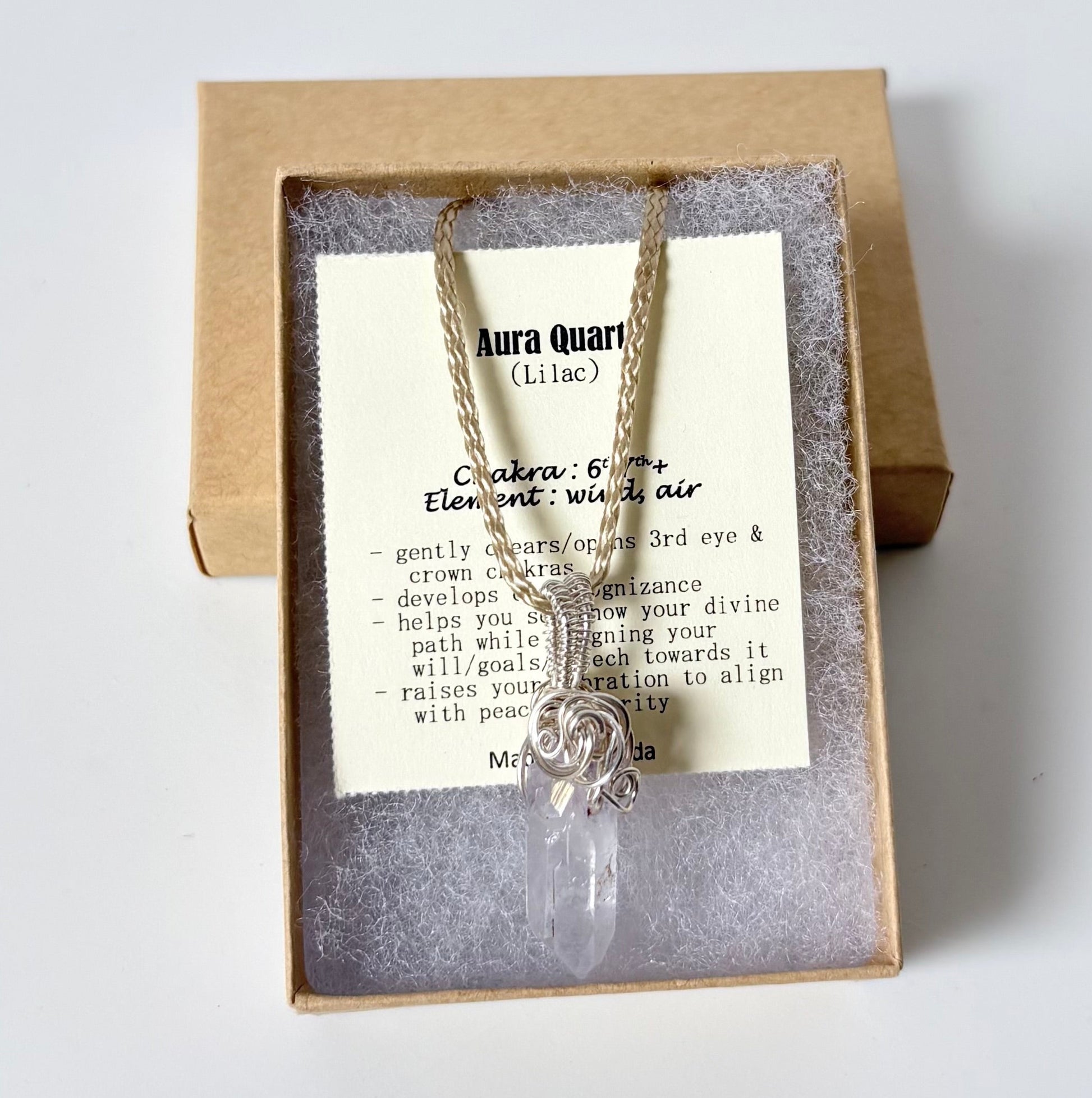 Necklace with a silver wire wrapped Aura Quartz (lilac) pendant on an information card with text explaining the folklore and metaphysical properties placed in a brown paper kraft jewelry box for gift giving all on a white surface.