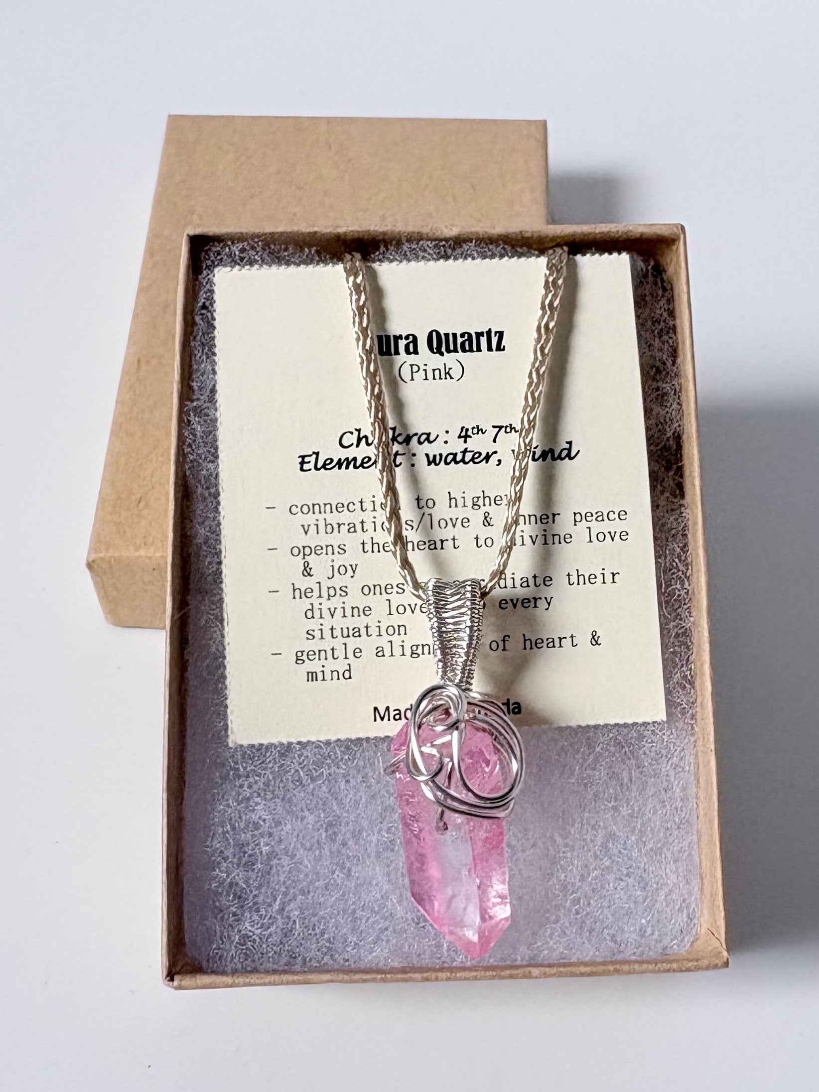 Necklace with a silver wire wrapped aura Quartz (pink) pendant on an information card with text explaining the folklore and metaphysical properties placed in a brown paper kraft jewelry box for gift giving all on a white surface.