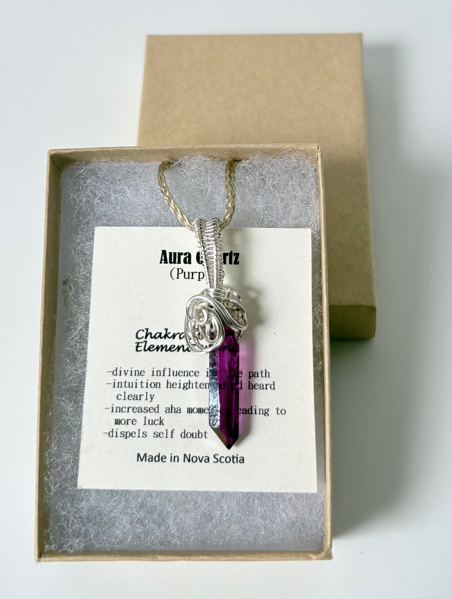 Necklace with a silver wire wrapped aura Quartz (purple) pendant on an information card with text explaining the folklore and metaphysical properties placed in a brown paper kraft jewelry box for gift giving all on a white surface.