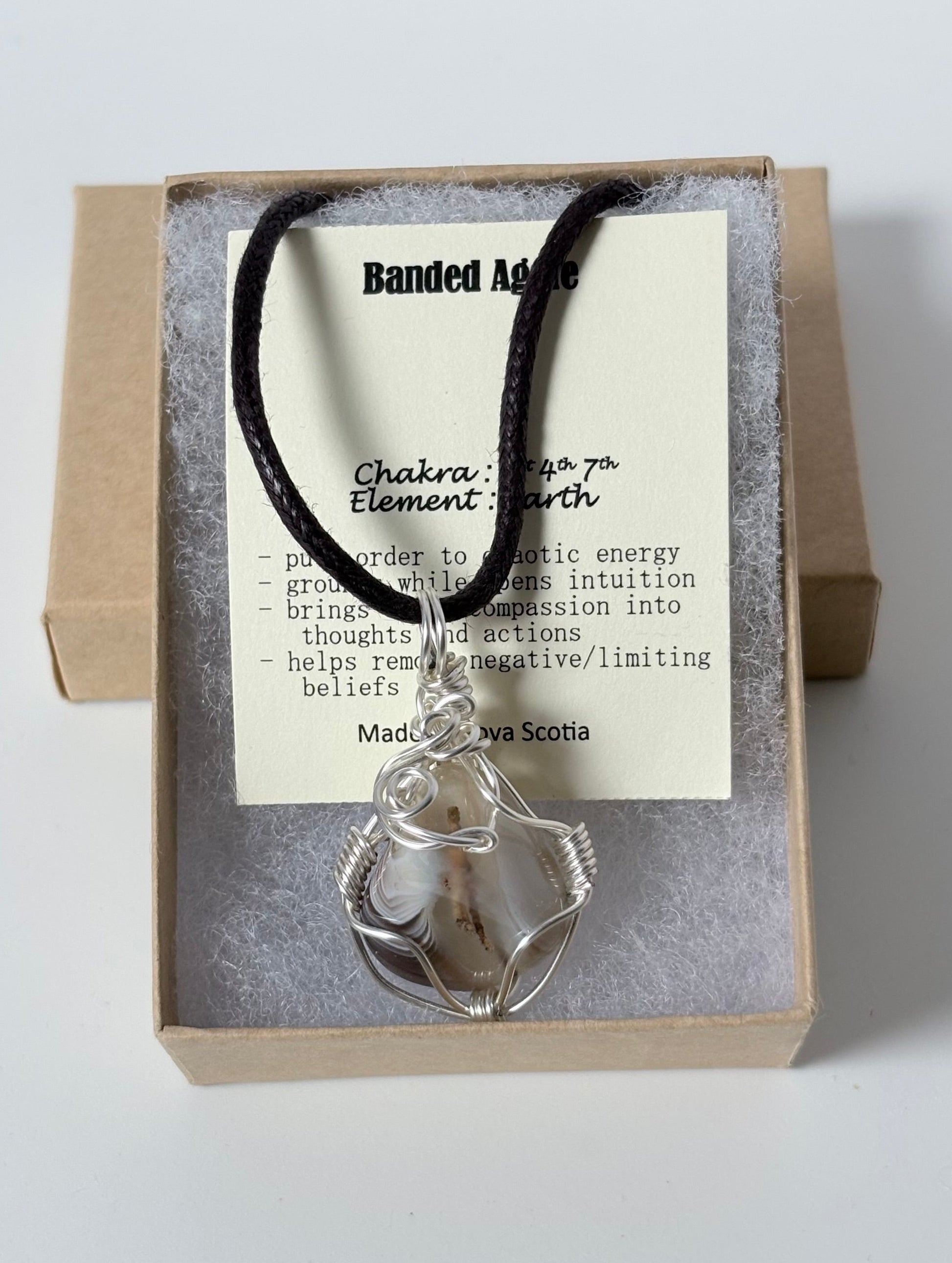 Necklace with a silver wire wrapped banded agate pendant on an information card with text explaining the folklore and metaphysical properties placed in a brown paper kraft jewelry box for gift giving all on a white surface.