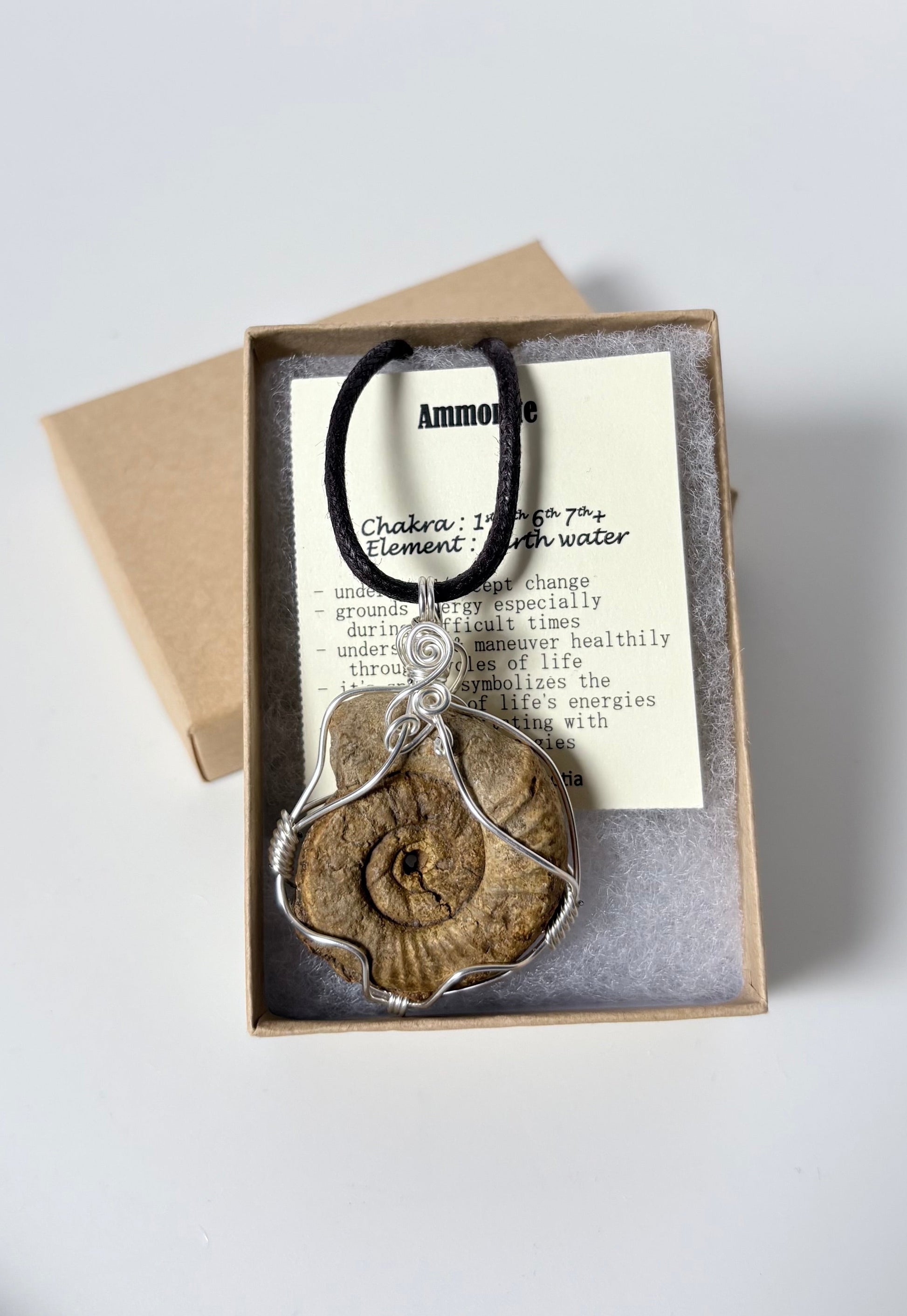 Necklace with a silver wire wrapped beige ammonite pendant on an information card with text explaining the folklore and metaphysical properties placed in a brown paper kraft jewelry box for gift giving all on a white surface.