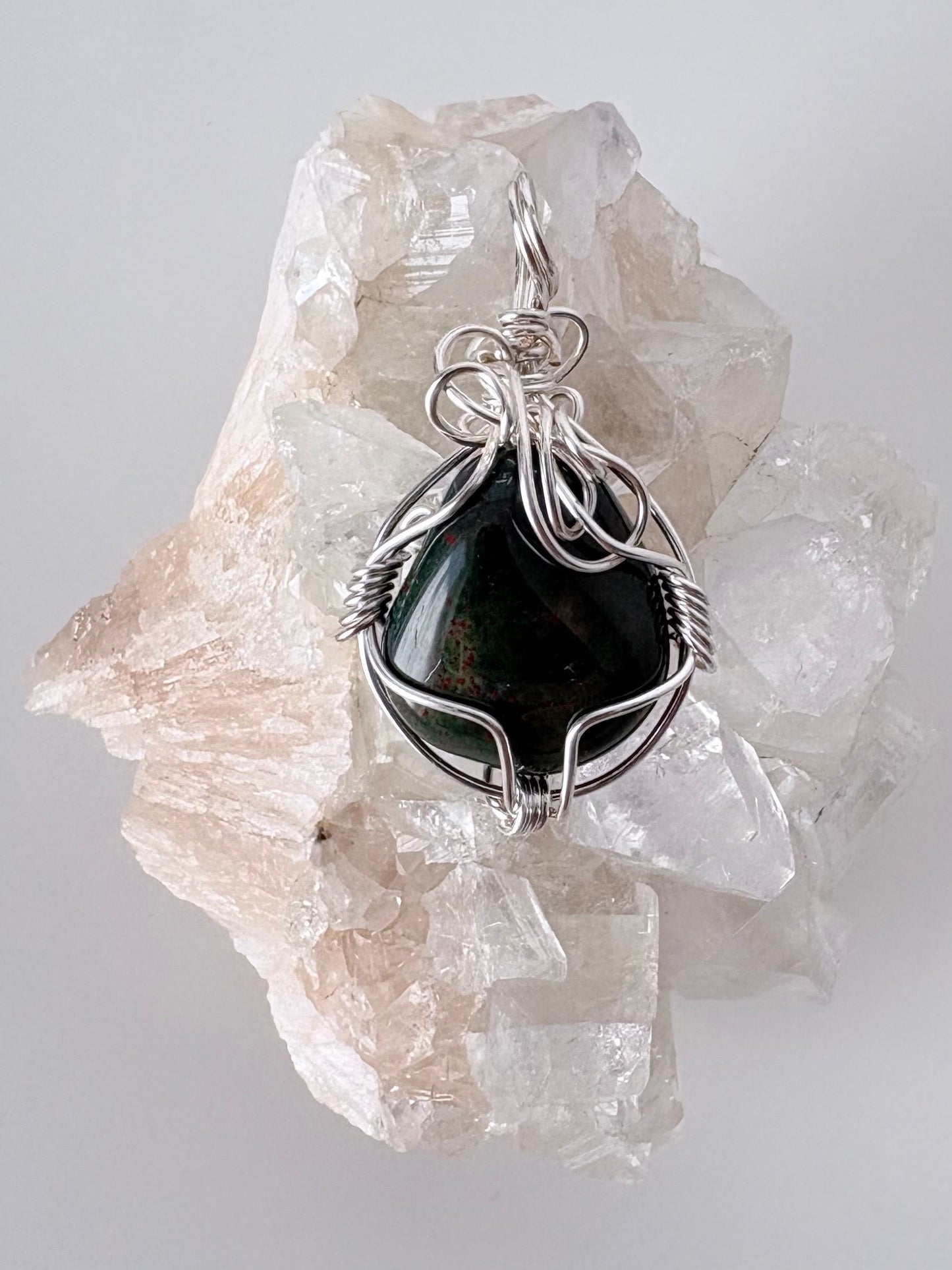 silver wire wrapped bloodstone pendant displayed on a clear apophyllite cluster all on a white surface.