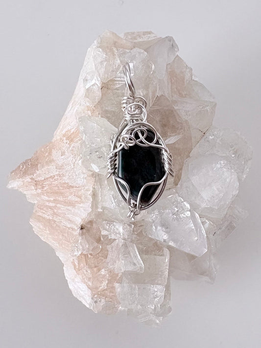 silver wire wrapped bloodstone pendant displayed on a clear apophyllite cluster all on a white surface.