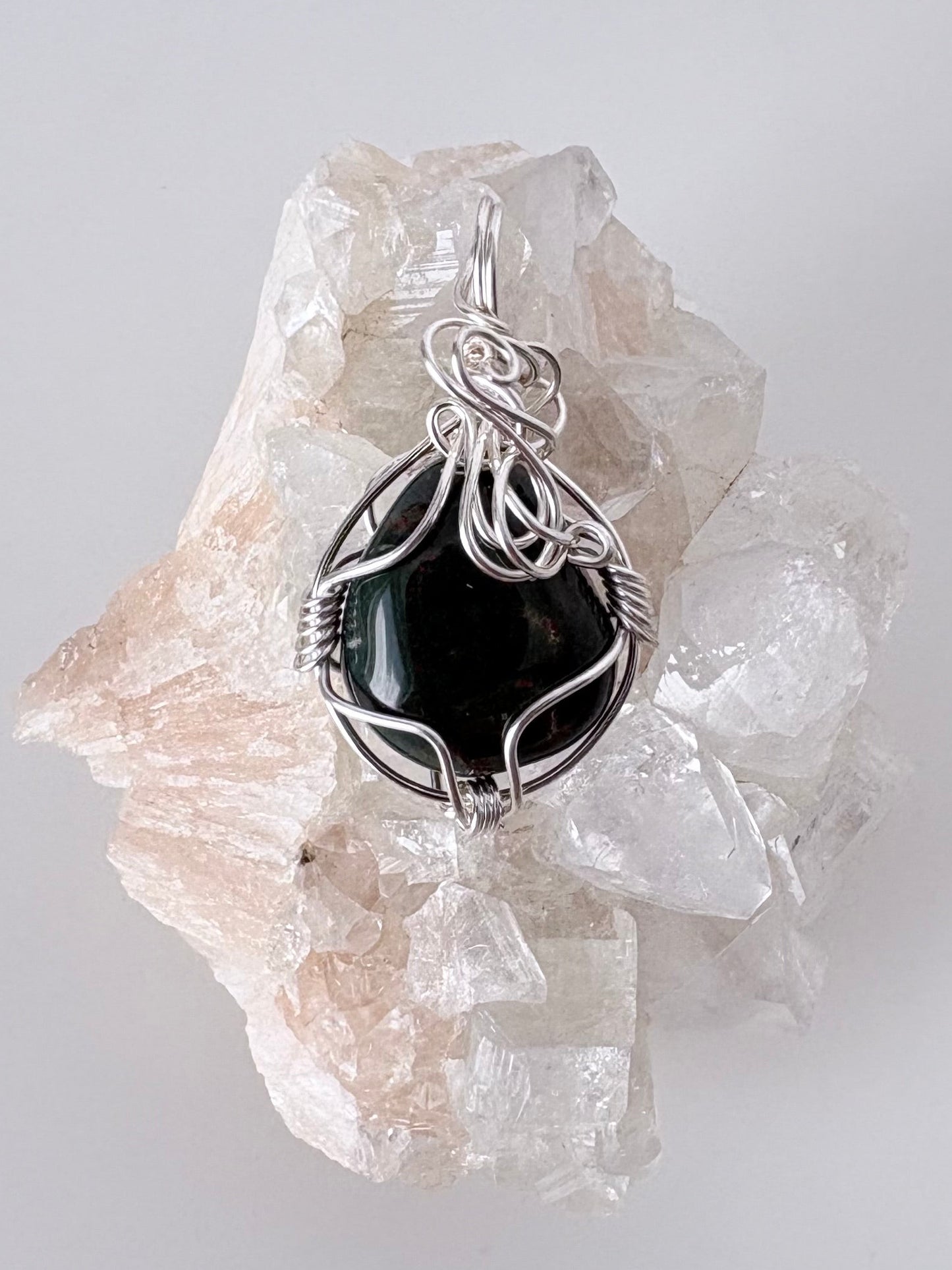 silver wire wrapped bloodstone pendant displayed on a clear apophyllite cluster all on a white surface.