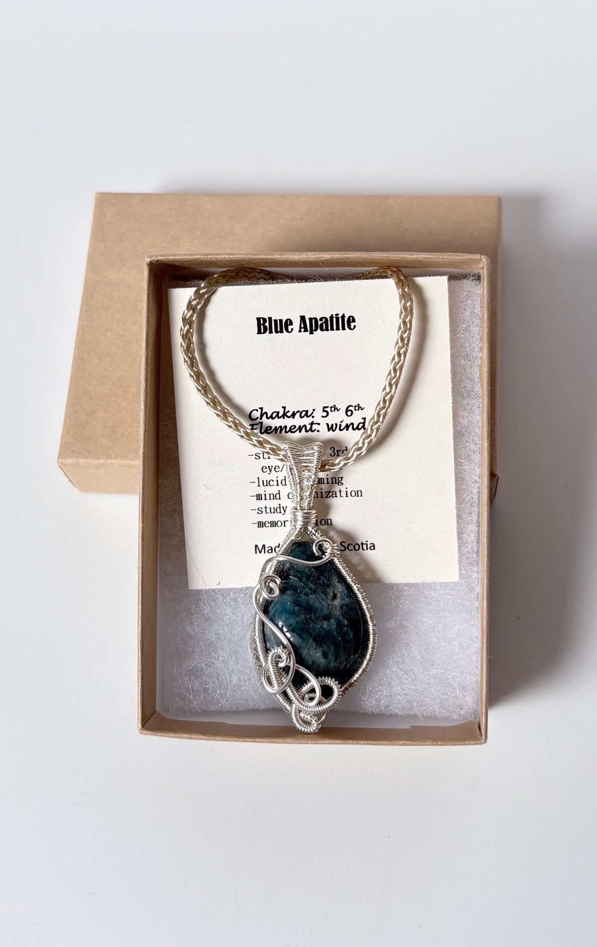 Necklace with a silver wire wrapped blue apatite pendant on an information card with text explaining the folklore and metaphysical properties placed in a brown paper kraft jewelry box for gift giving all on a white surface.