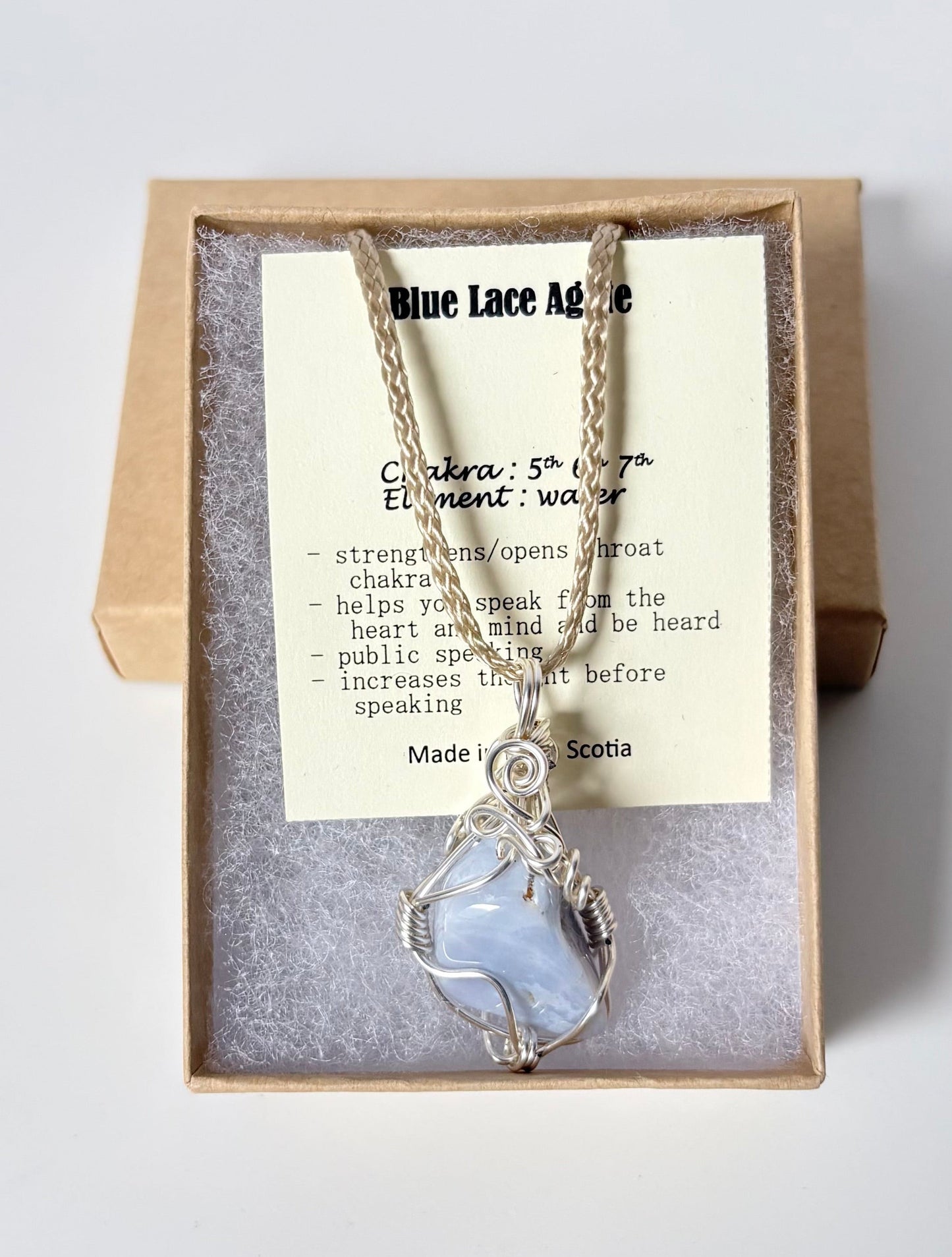 Necklace with a silver wire wrapped blue lace agate pendant on an information card with text explaining the folklore and metaphysical properties placed in a brown paper kraft jewelry box for gift giving all on a white surface.