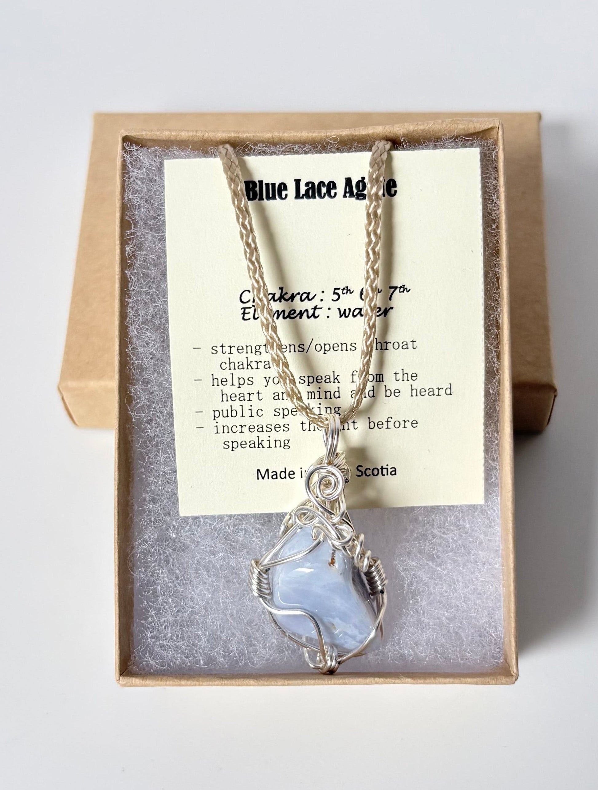Necklace with a silver wire wrapped blue lace agate pendant on an information card with text explaining the folklore and metaphysical properties placed in a brown paper kraft jewelry box for gift giving all on a white surface.