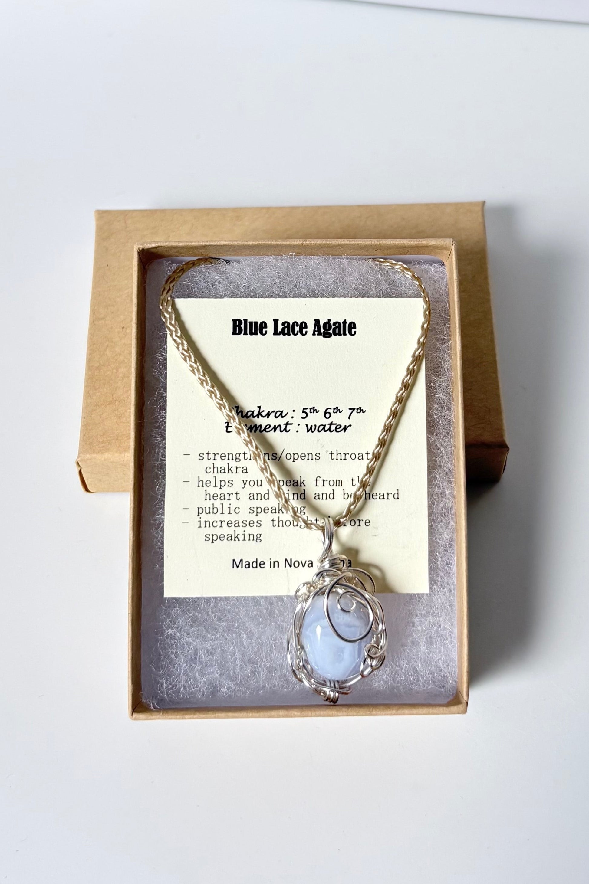 Necklace with a silver wire wrapped blue lace agate pendant on an information card with text explaining the folklore and metaphysical properties placed in a brown paper kraft jewelry box for gift giving all on a white surface.
